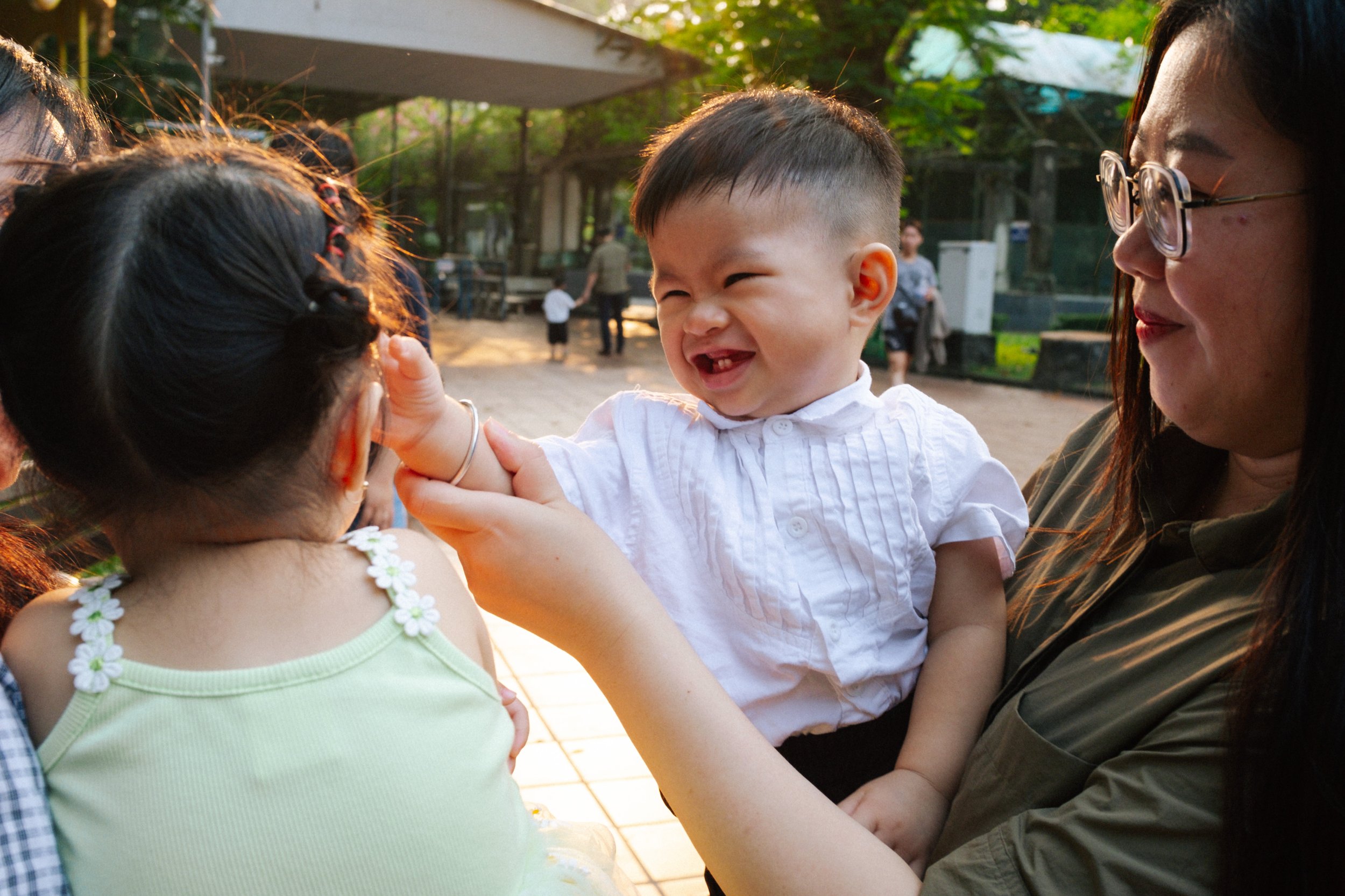 A young boy smiling and laughing while being held by a woman wearing glasses, as a girl with braided hair and a yellow dress looks at him. People are in the background outdoors during sunset.
