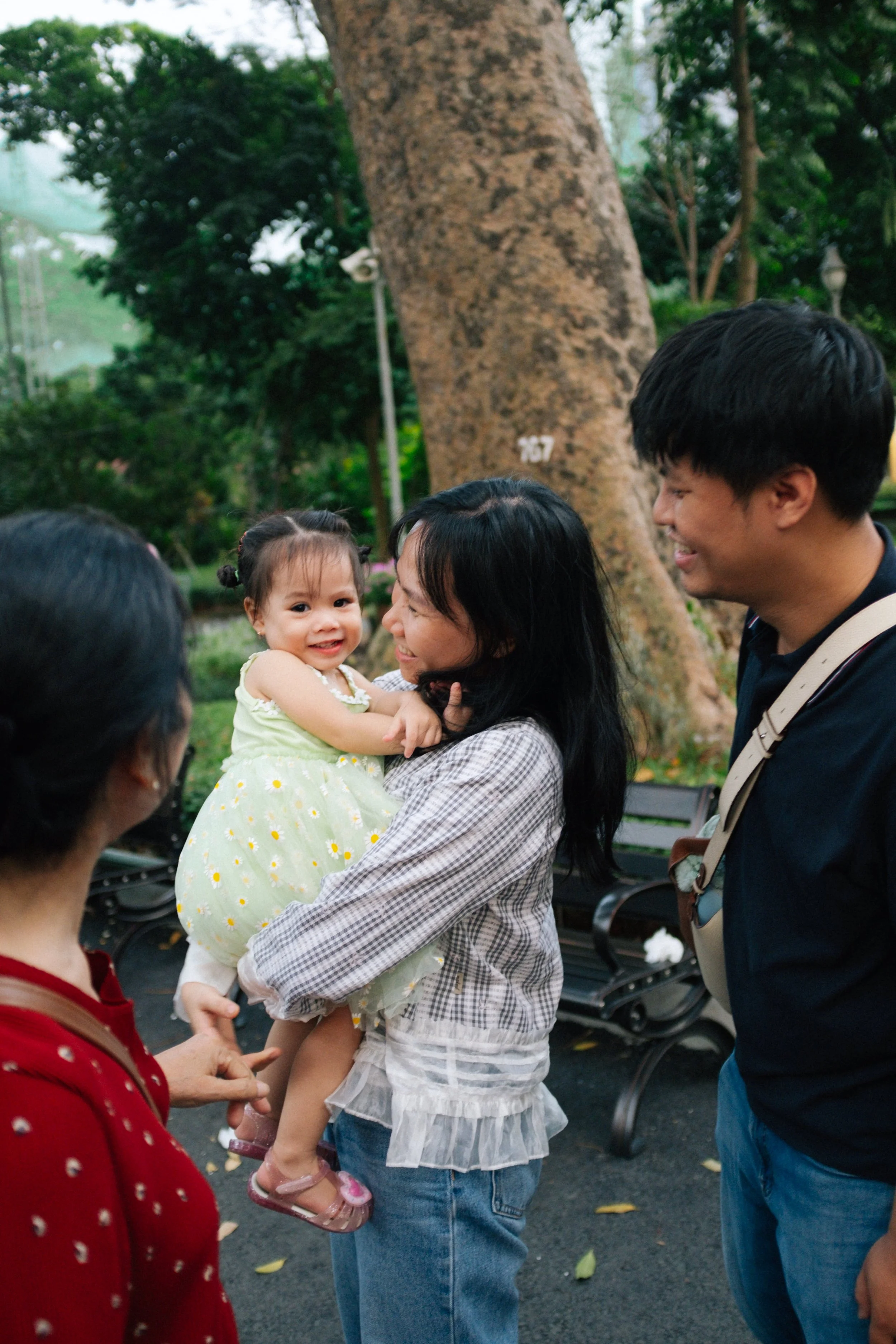 A woman holding a young girl in a park, smiling. A man and another woman are nearby, also smiling. The background shows trees and park benches.