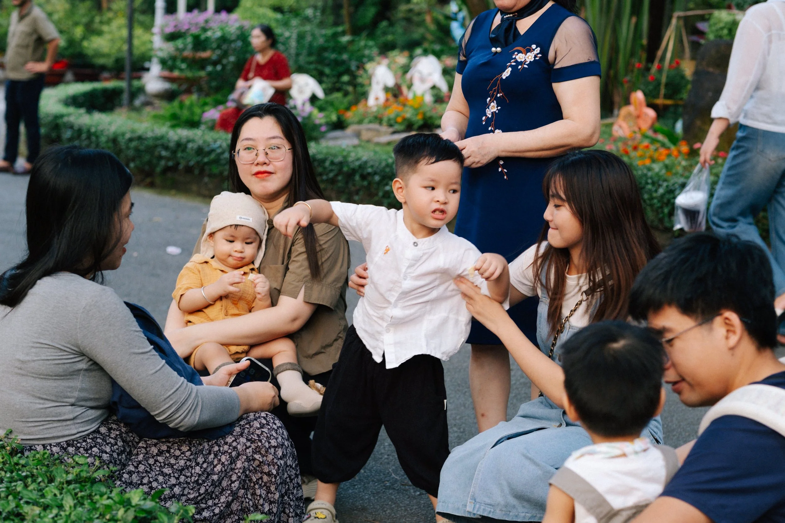 Group of Asian women and children gathered outdoors in a park, with some sitting and others standing, surrounded by green plants and flowers.