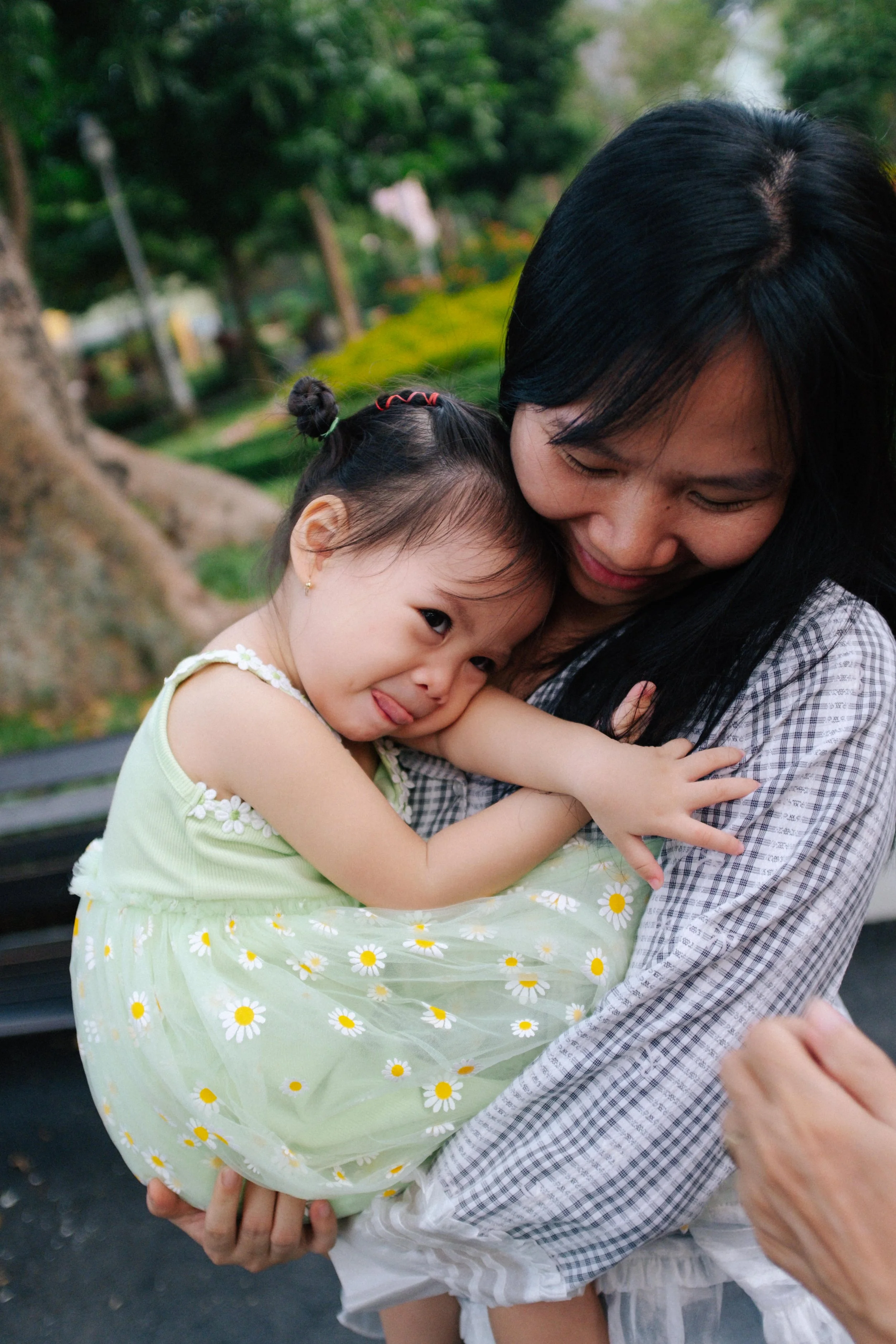 A woman holding a girl dressed in a yellow and white floral dress, sharing a tender embrace outdoors in a park with trees and green foliage.