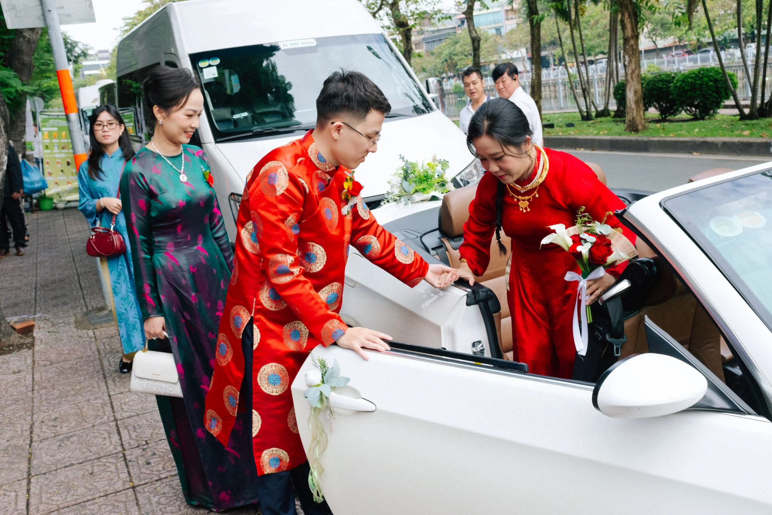 People dressed in traditional Vietnamese clothing, Ao Dai, participating in a wedding ceremony with a car decorated with flowers.