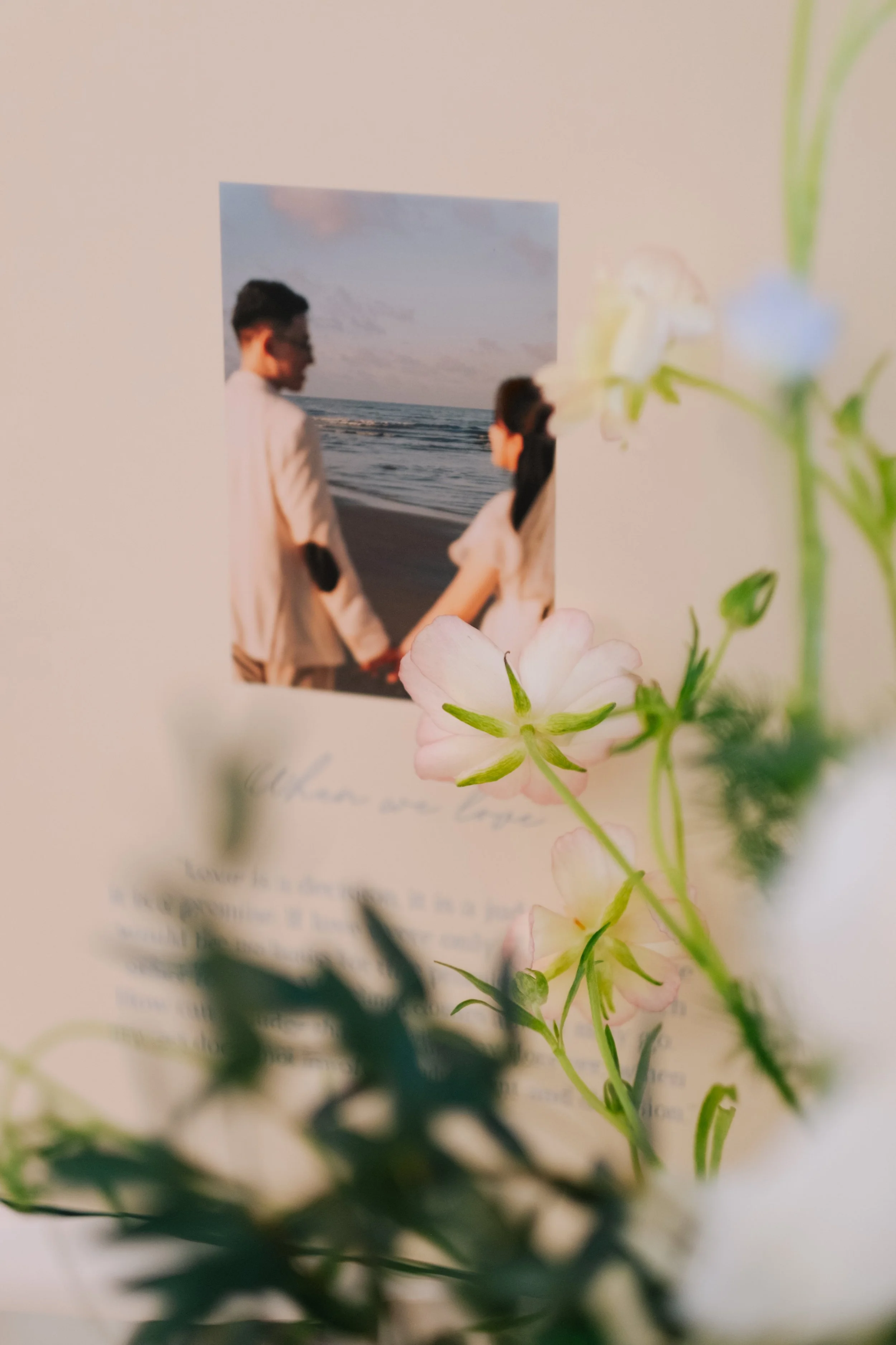 A photo of a couple holding hands on the beach at sunset, viewed through pink and white flowers with some text at the bottom.