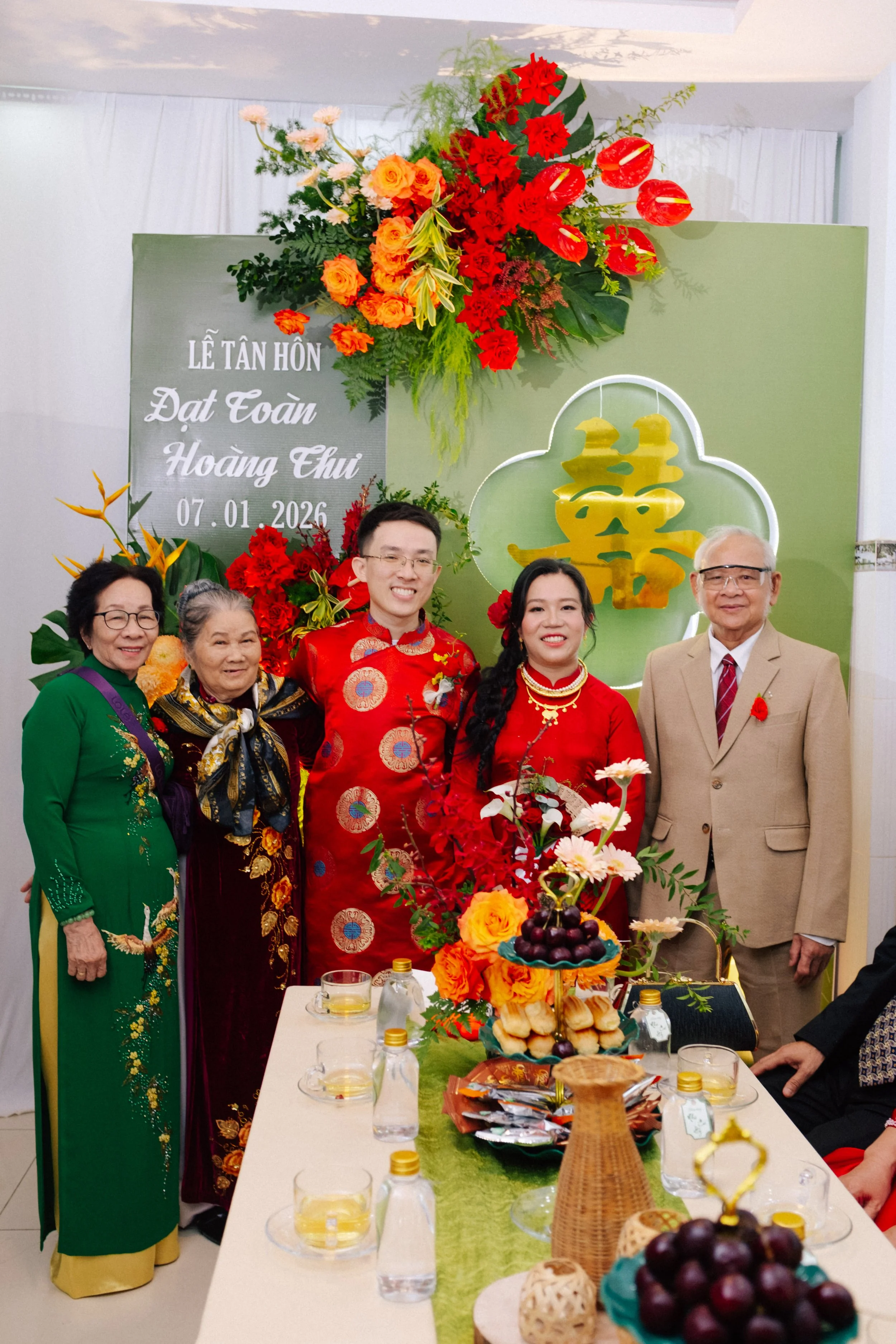 A family celebrating a wedding in Vietnam, with a decorated table, floral arrangements, and a green backdrop with gold Chinese character for 'longevity' and wedding date.