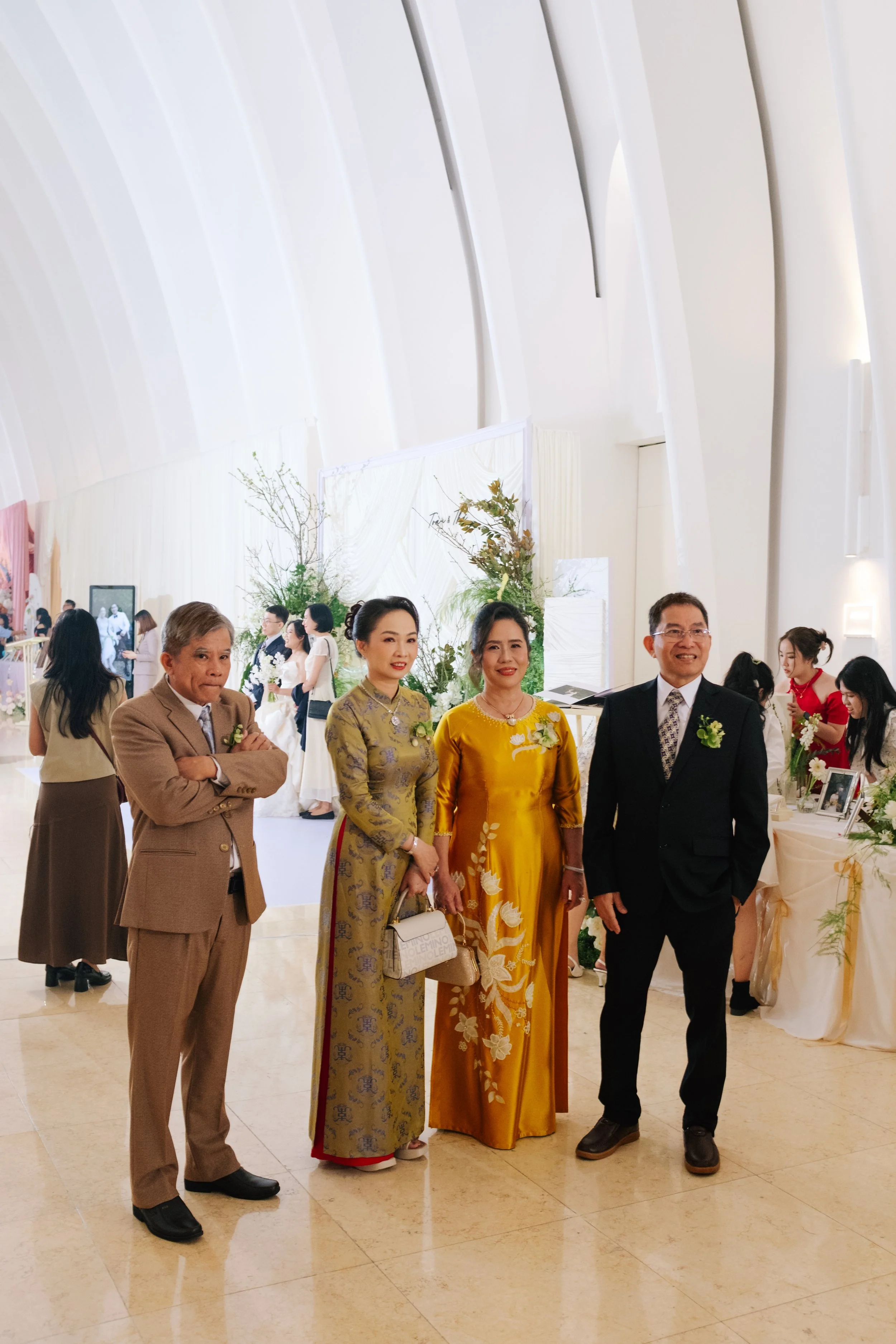 Group of four people dressed in formal Southeast Asian attire at a wedding reception, standing on a light-colored tiled floor with a white, curved wall in the background and wedding decorations.