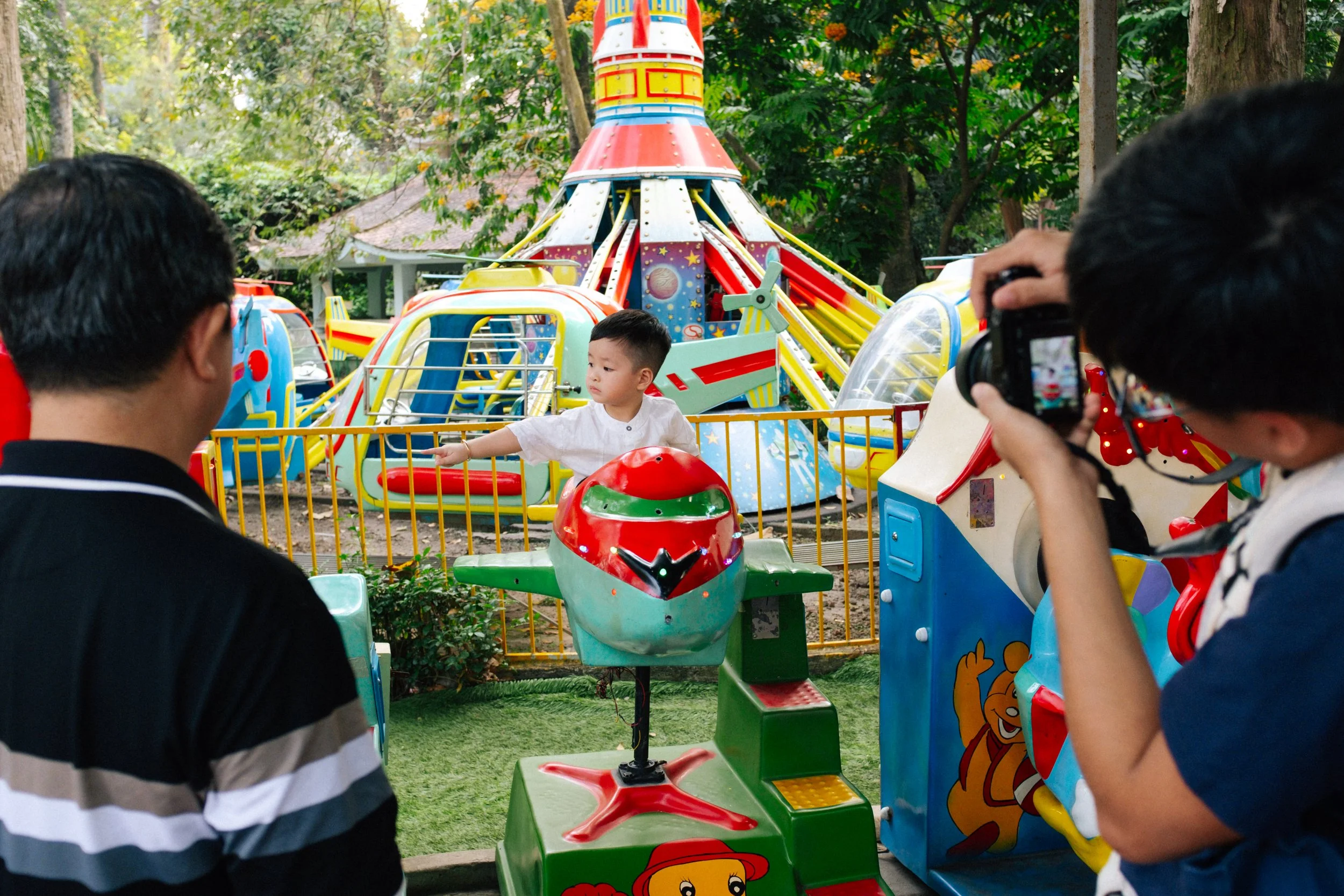 A young boy at an amusement park or carnival, with two adults taking photos of him, as a kiddie ride featuring a spaceship and a small carousel horse are visible in the background, surrounded by trees and colorful amusement rides.