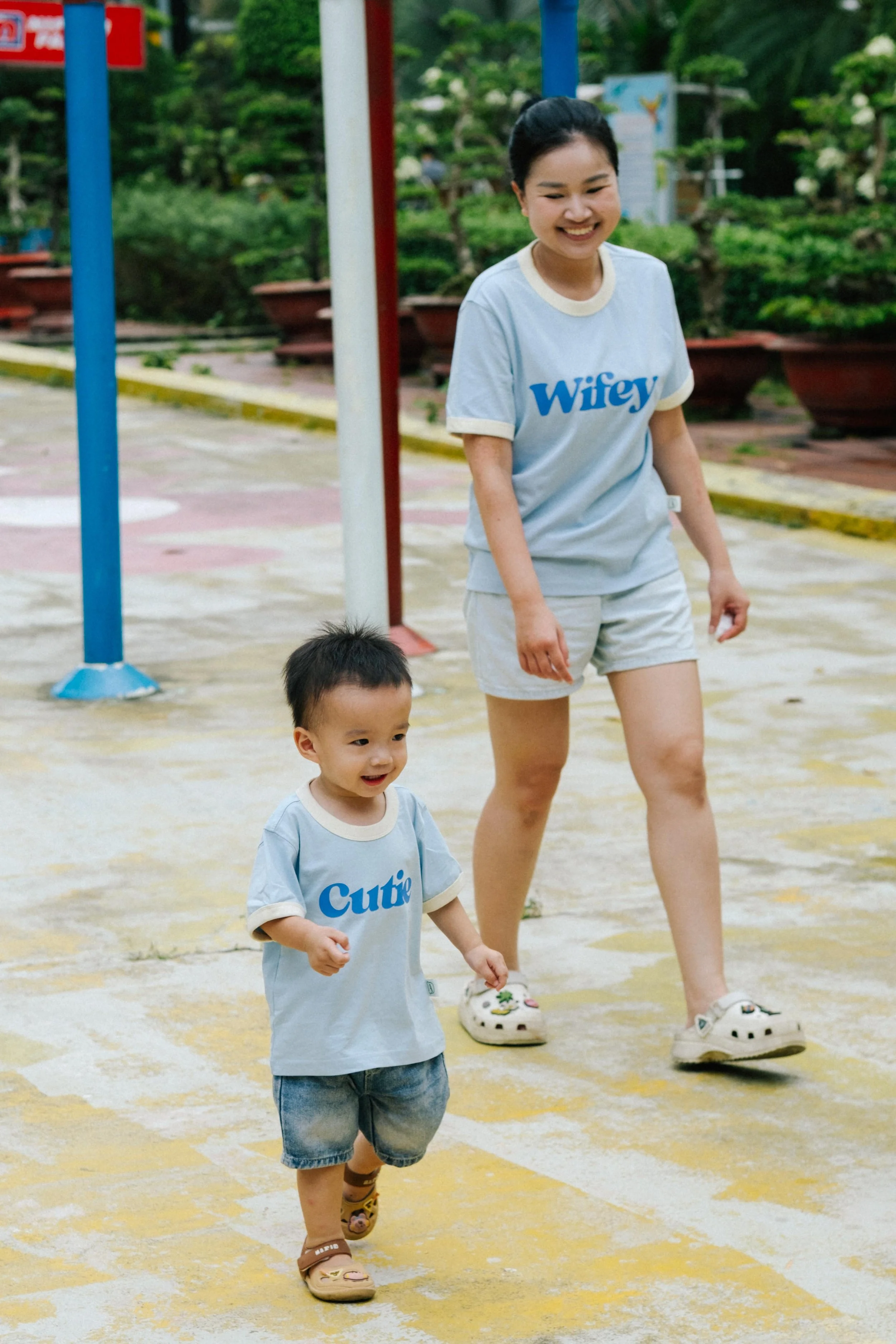 A woman and a young boy walking outdoors on a colorful concrete surface, smiling and wearing matching light blue T-shirts with words 'Wifey' and 'Cutie' printed on them.