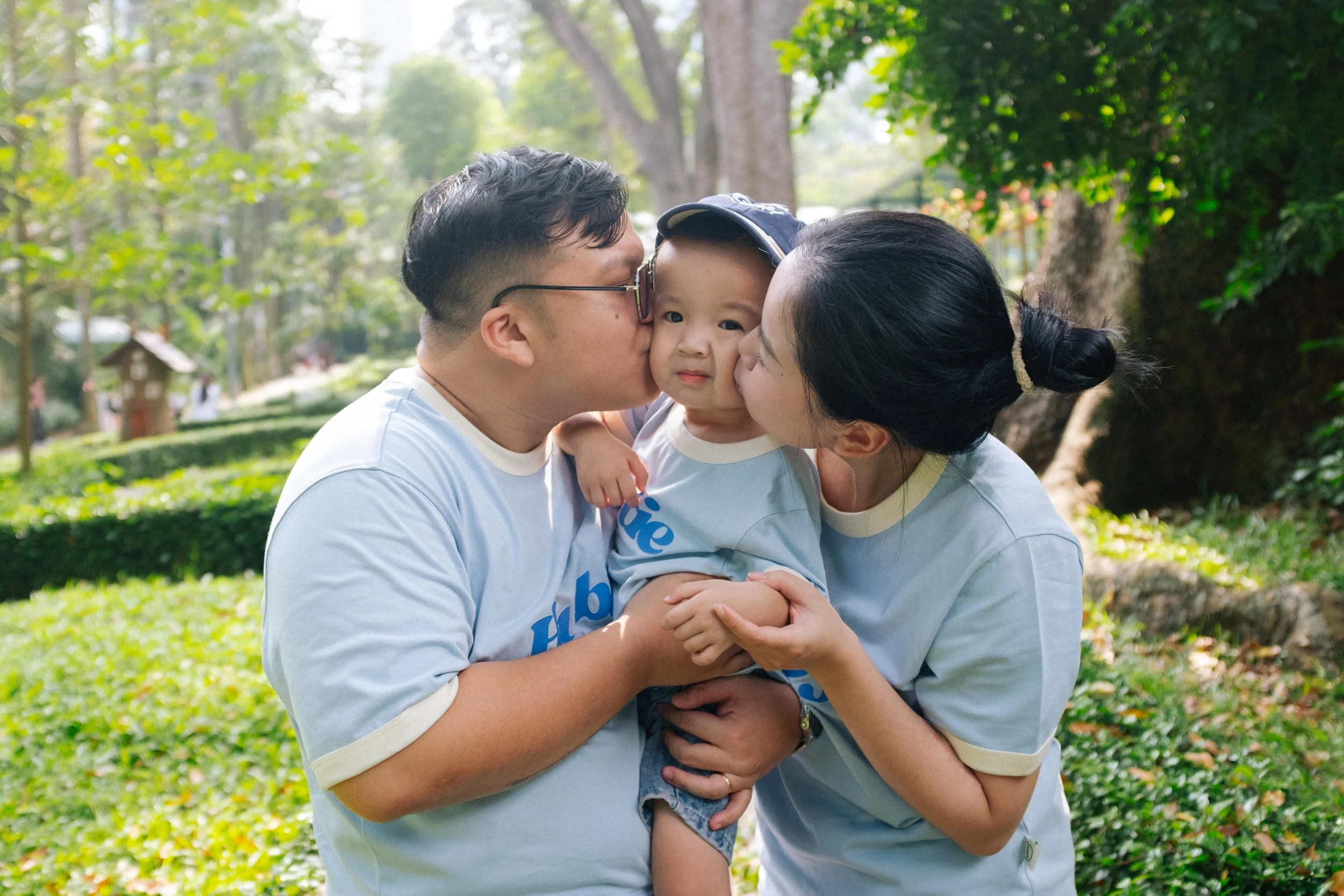 A family of three outdoors in a park, with the father and mother kissing their young son on the cheeks. The family is wearing matching light-colored shirts, standing in front of green trees and bushes.