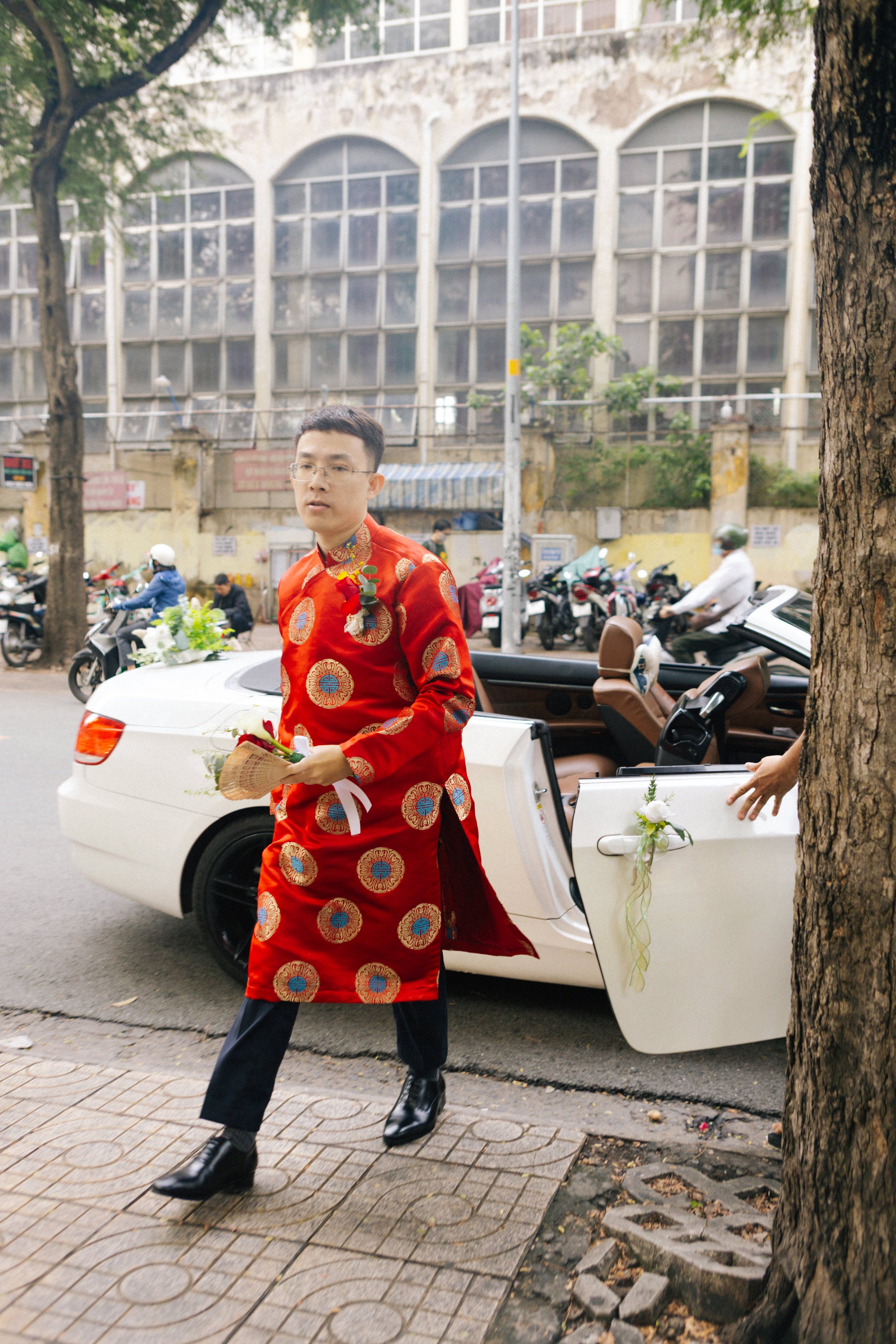 A man in traditional red wedding attire walking on the sidewalk, holding a fan and wearing a boutonniere, after a wedding ceremony. A white convertible car decorated with flowers is parked behind him.