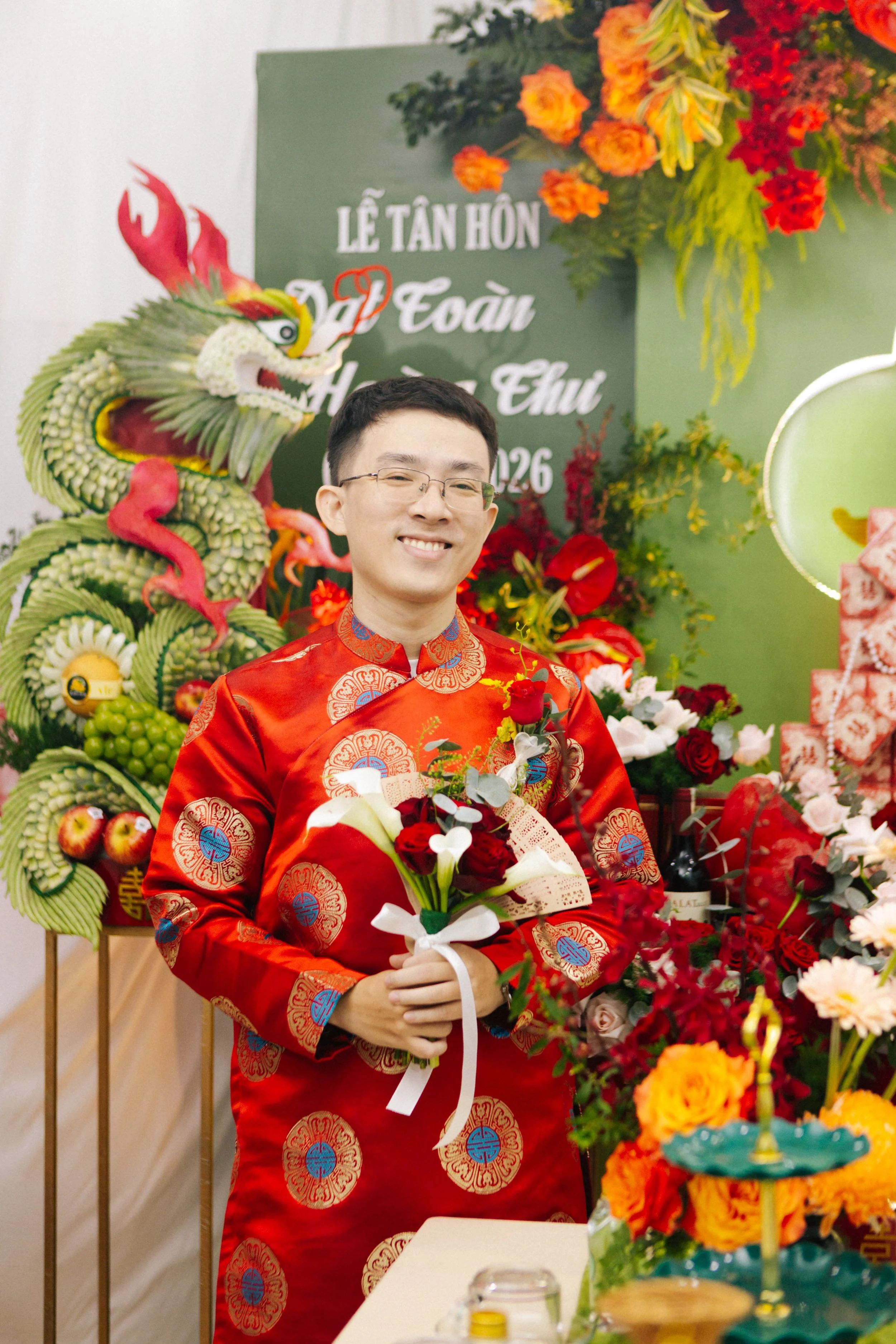 A man dressed in a traditional red Asian wedding outfit holding a bouquet of flowers, smiling at a wedding celebration with colorful floral arrangements and a dragon sculpture in the background.