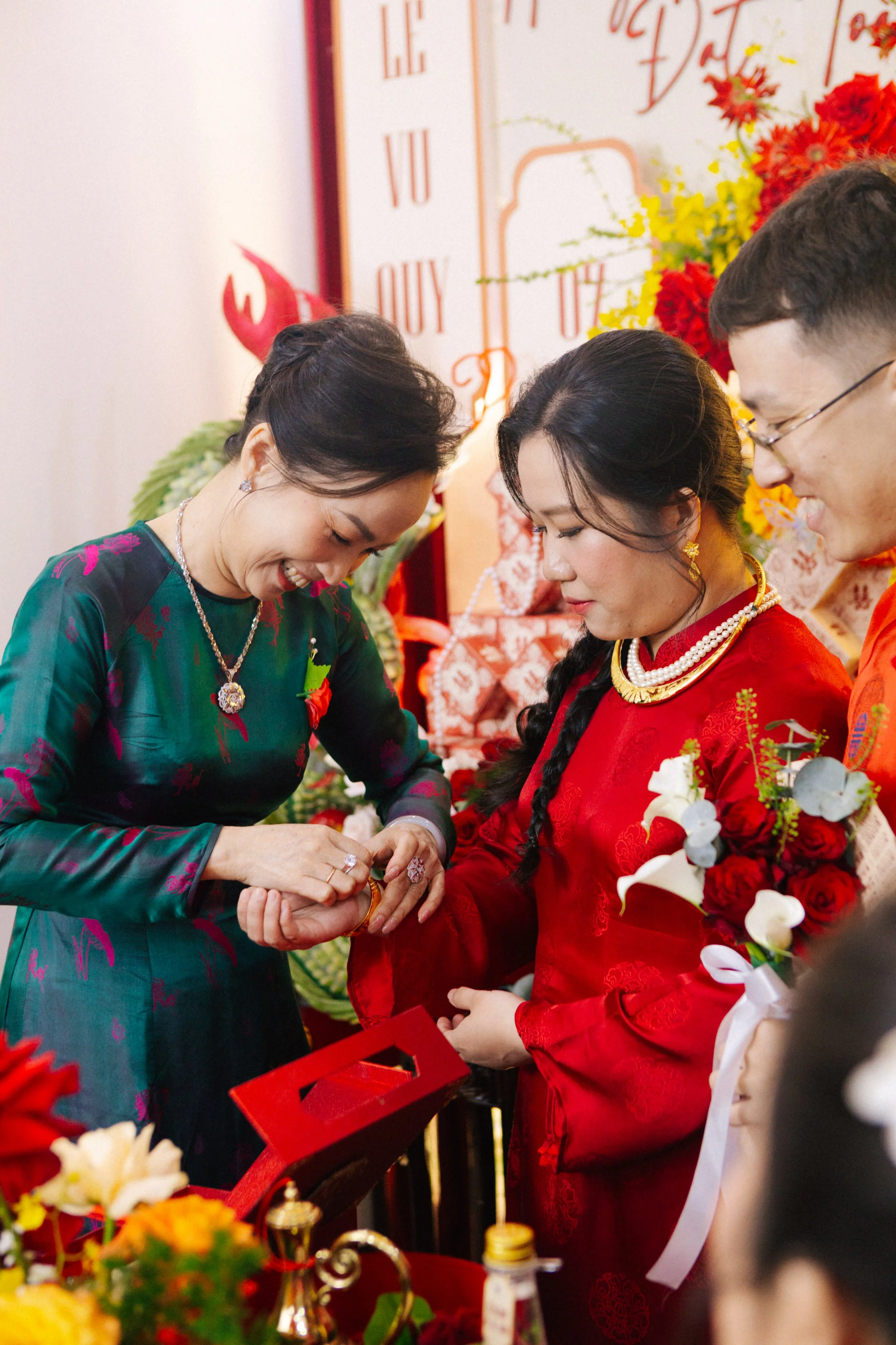 A wedding ceremony where a bride and groom are exchanging rings, with an officiant assisting. The bride is dressed in a red traditional outfit, holding a bouquet of red and white flowers, and other guests are present.