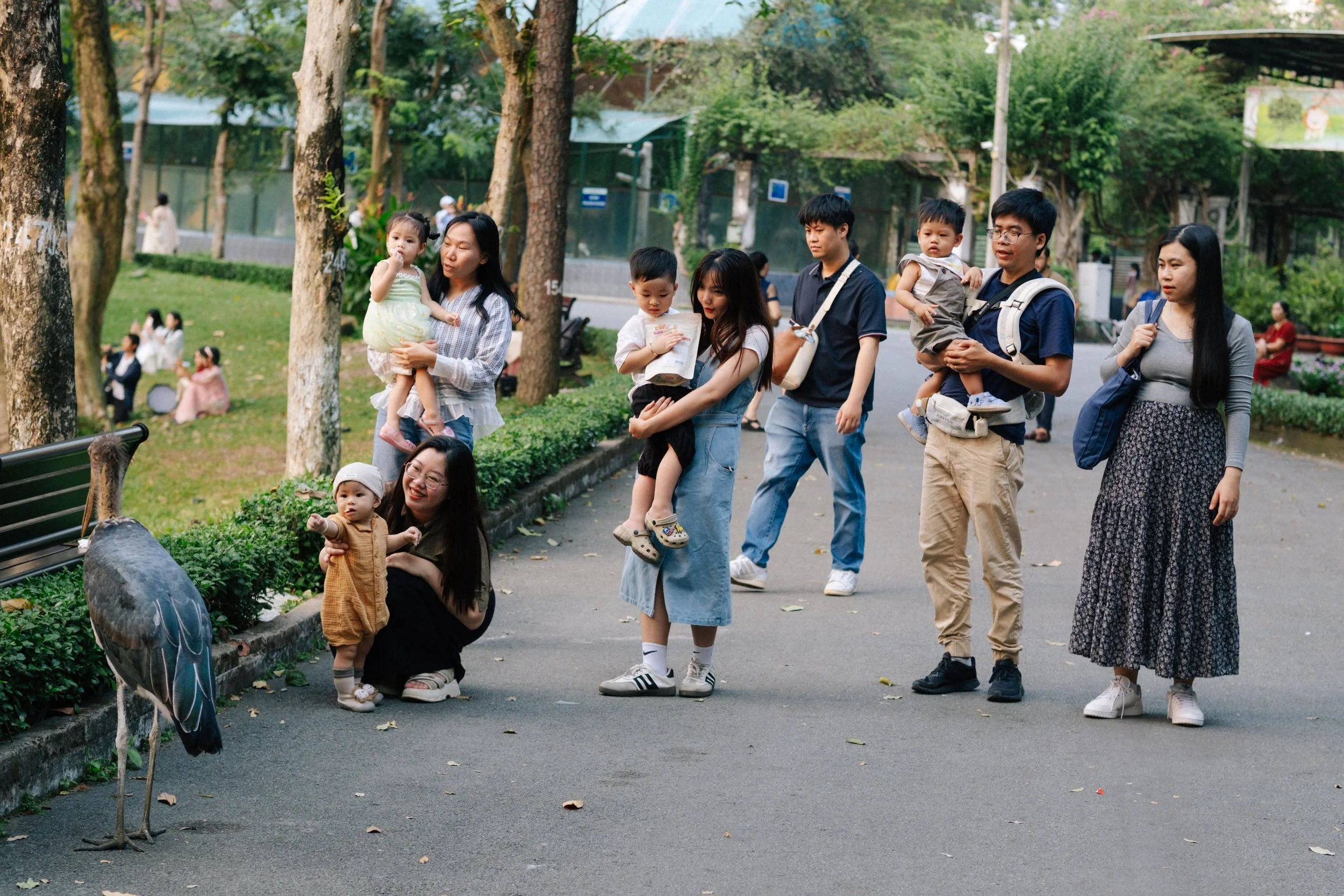 Group of people, including children and adults, observing a large bird near a park pathway with trees and greenery in the background.