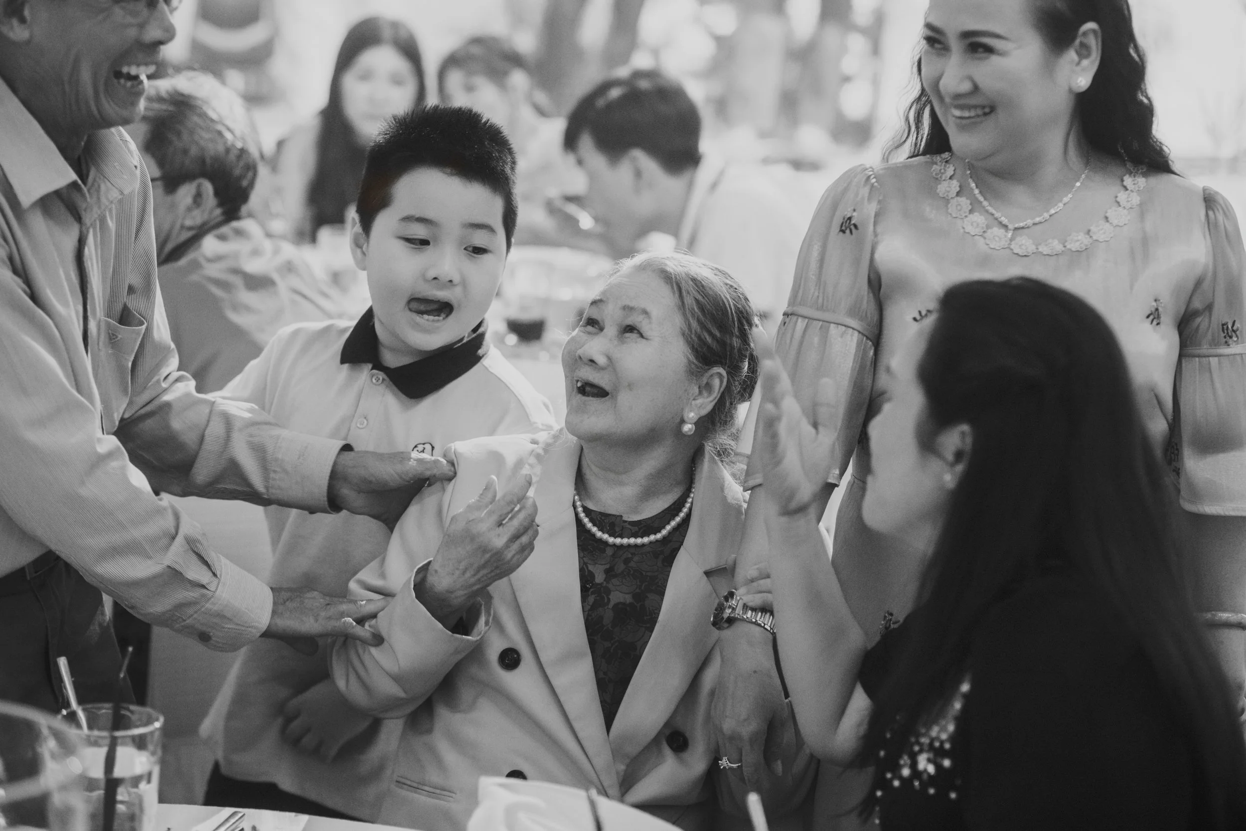 People enjoying a joyful gathering outdoors, with an elderly woman in the center smiling and a young boy talking animatedly, surrounded by adults, in a festive atmosphere.