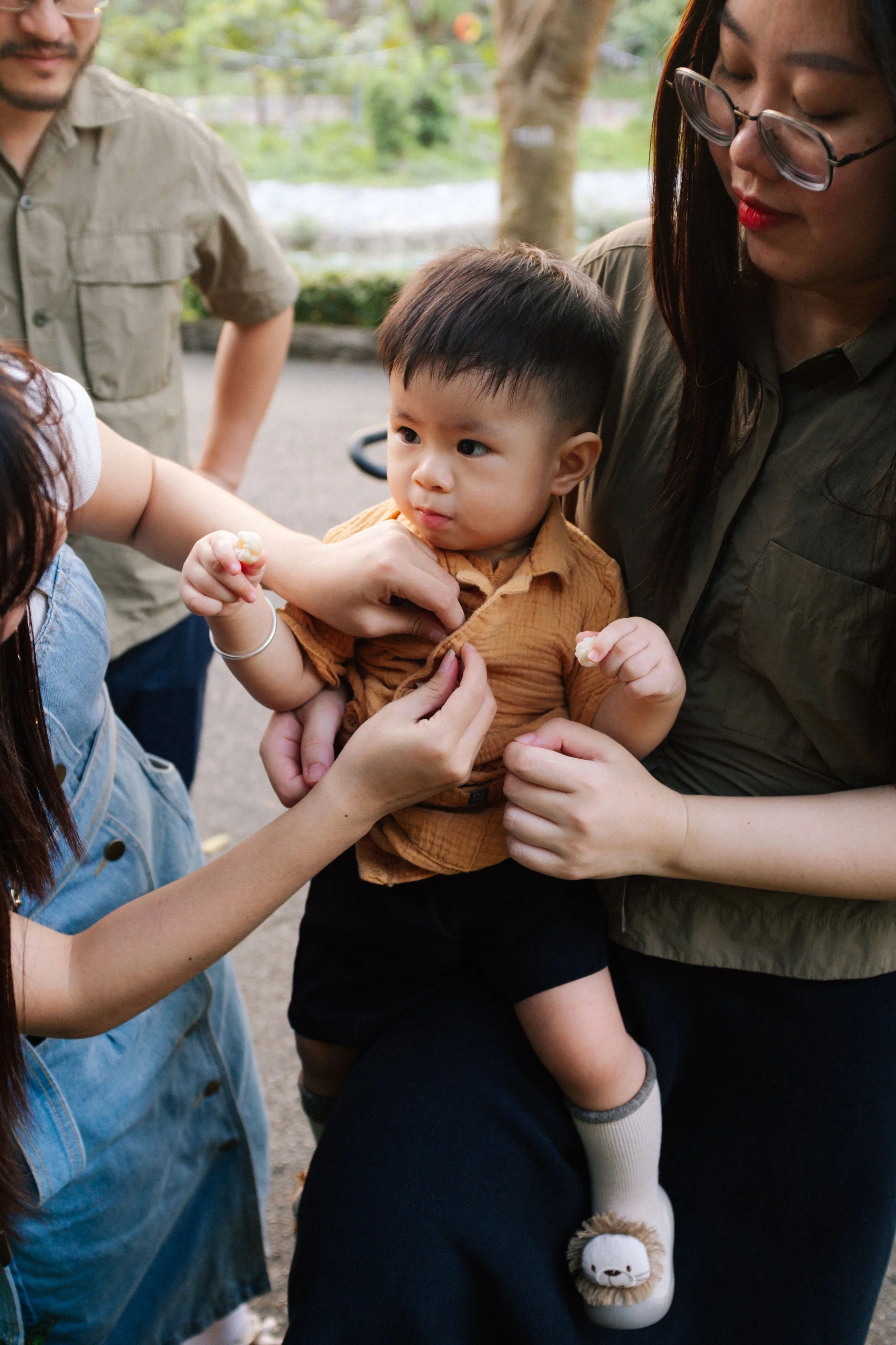 A young Asian boy in an orange shirt and black shorts being held by a woman, while another woman assists, outside in a park-like setting.
