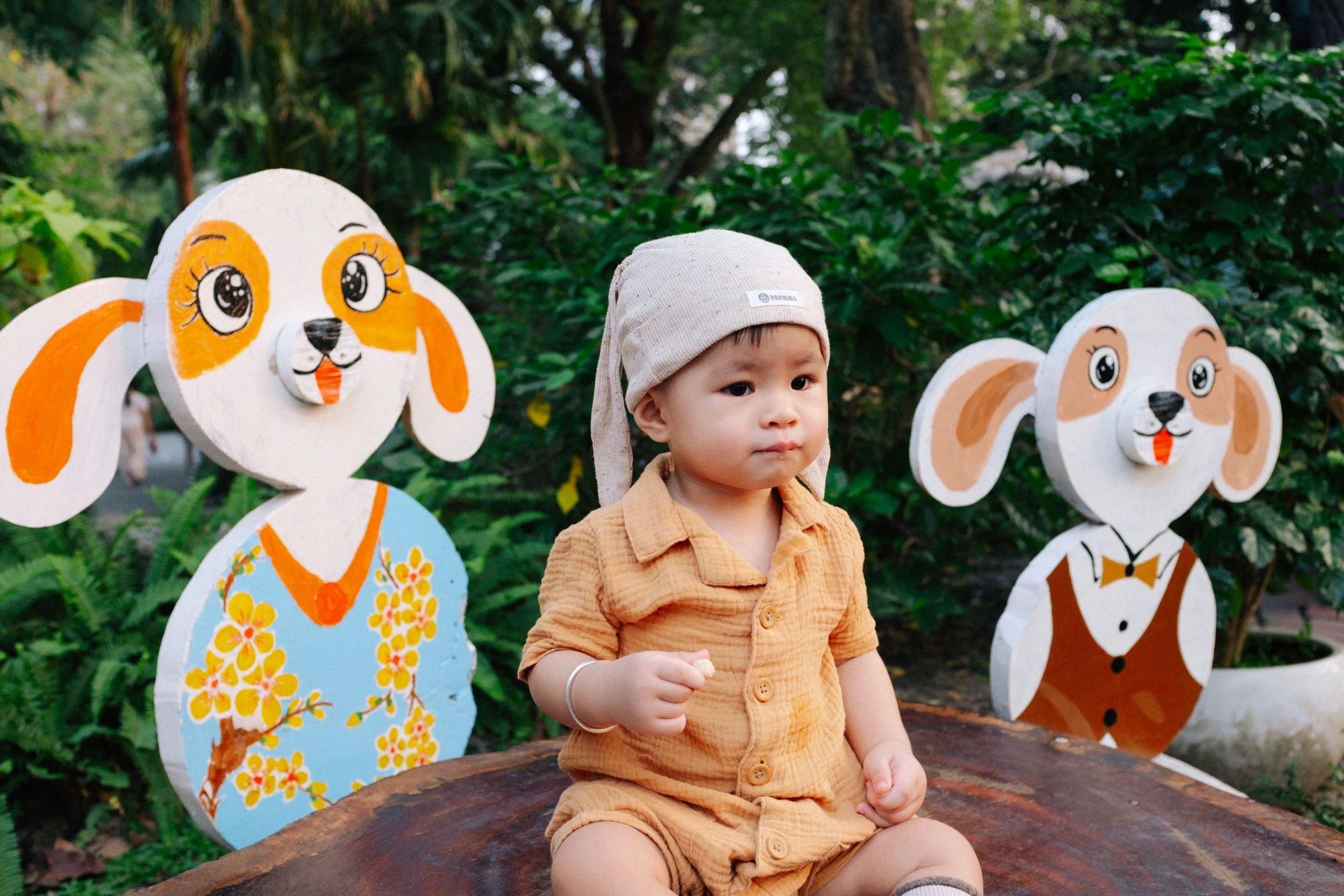 Young boy sitting on a wooden surface outdoors, wearing a beige outfit and a beige headscarf, with two cartoon dog cutouts in the background among green foliage.