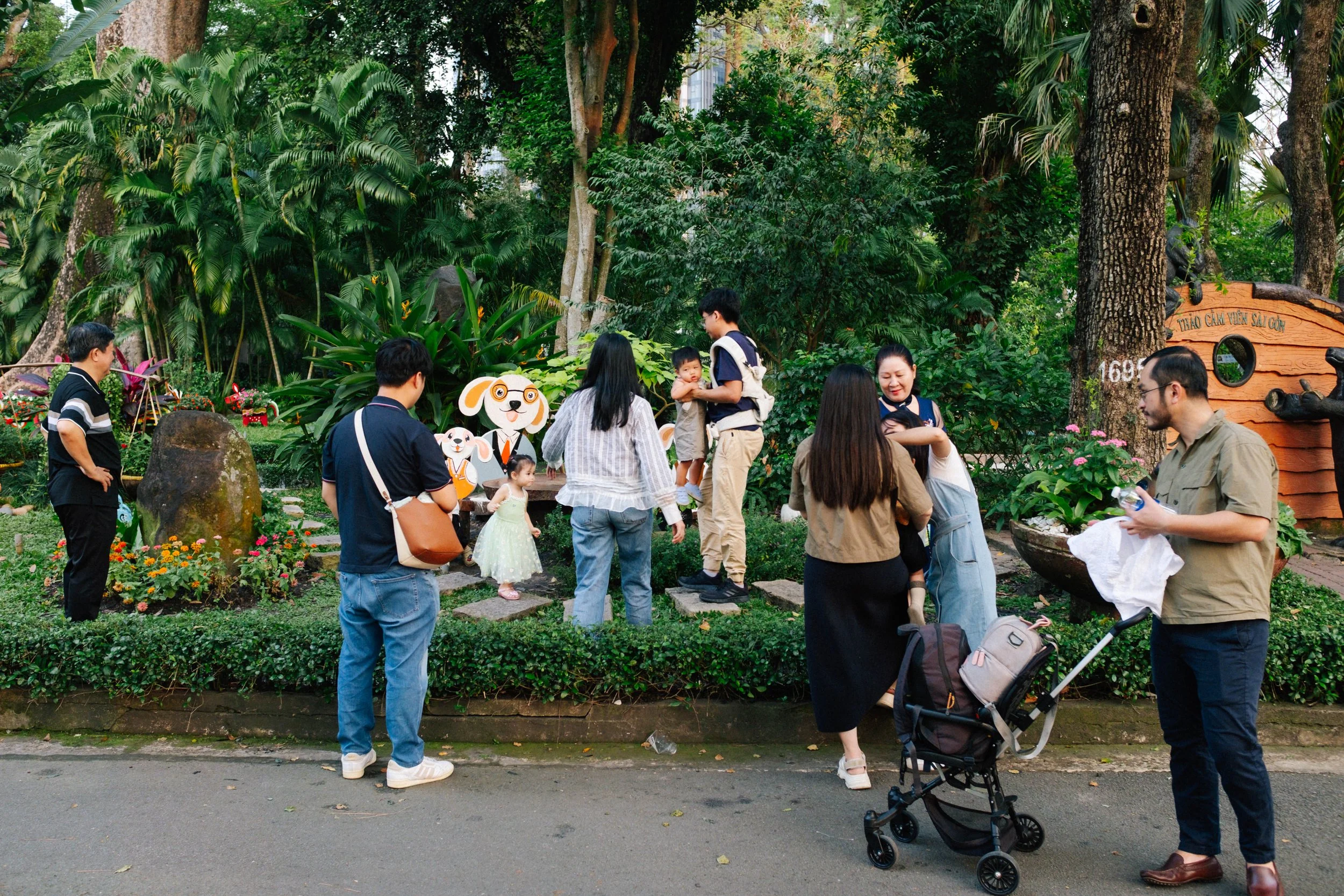 People gathering in a garden with children and a dog-themed cartoon cutout, surrounded by trees and plants.