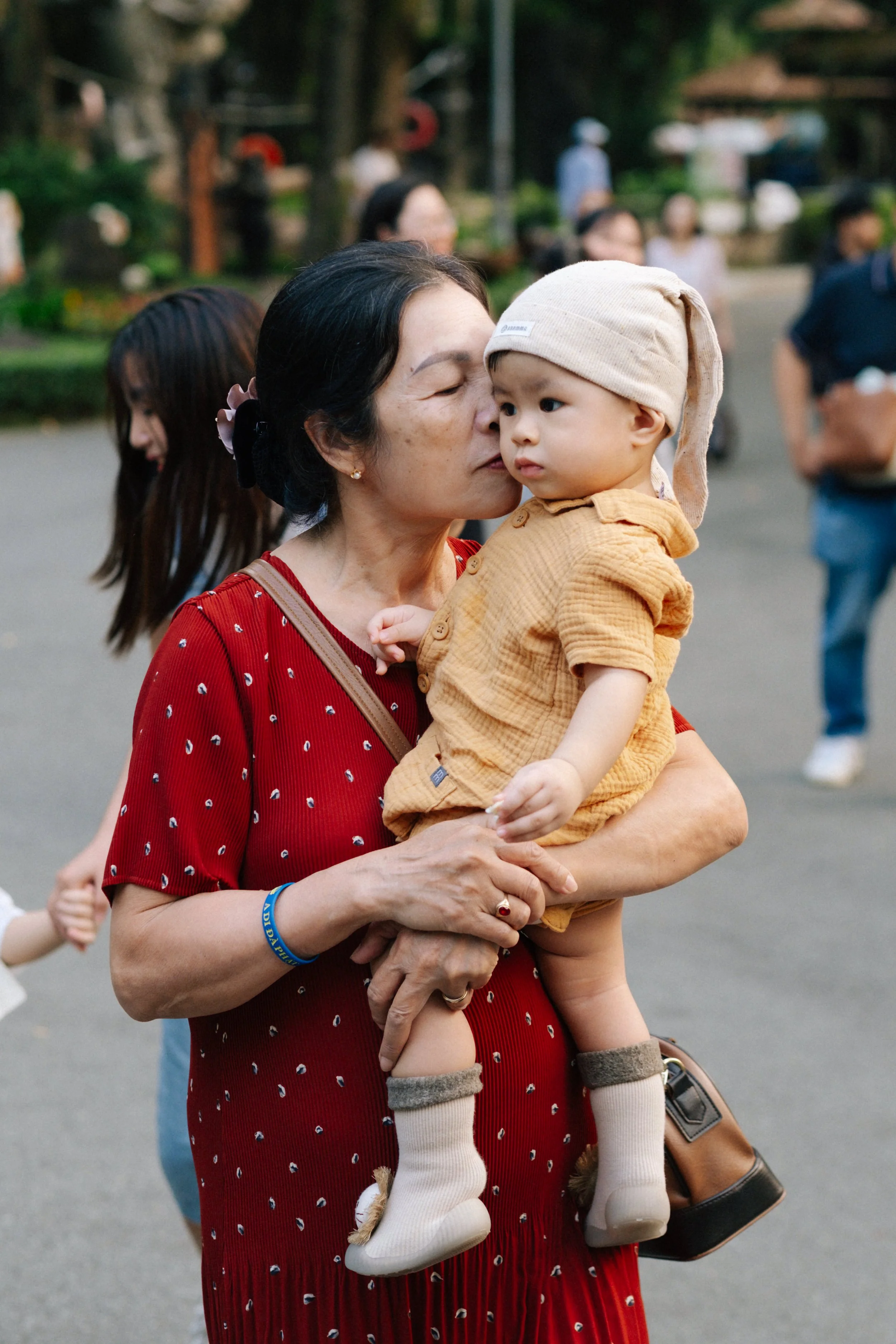 A woman in a red dress holding a young child wearing a beige hat and orange outfit, giving a gentle kiss on the child's face.