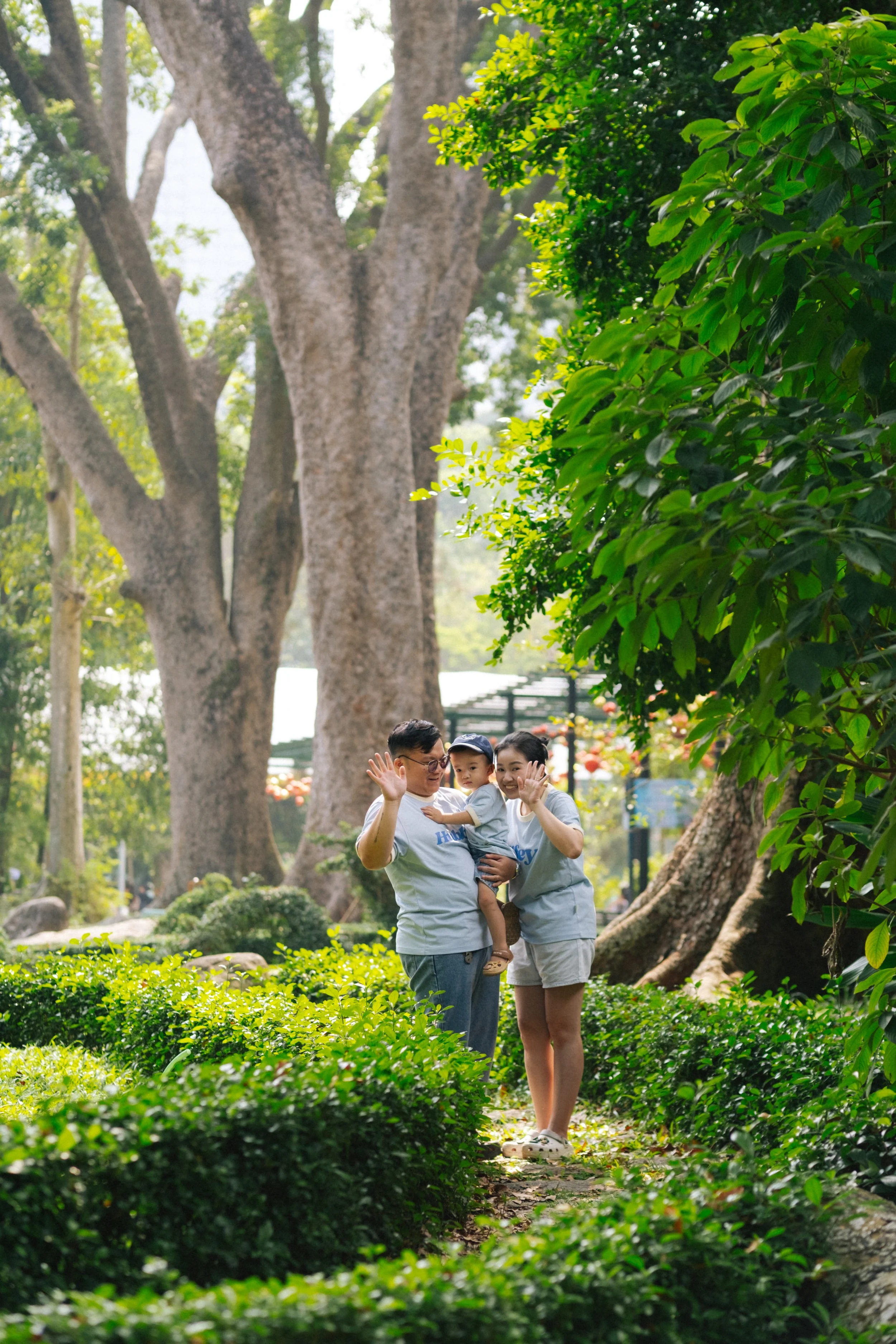 A family of three in a park with large trees and lush green foliage, waving at the camera.
