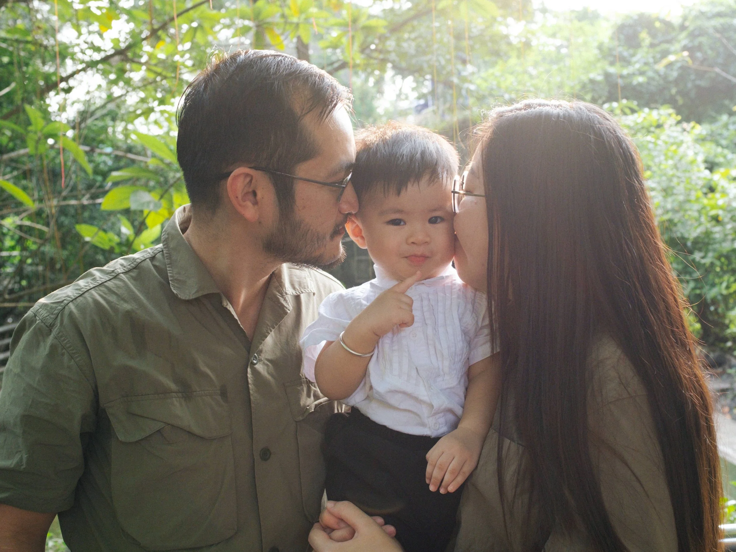 A family of three, a man, woman, and young boy, sharing a loving moment outdoors, with the woman and man kissing the boy on each cheek in a green, natural setting.