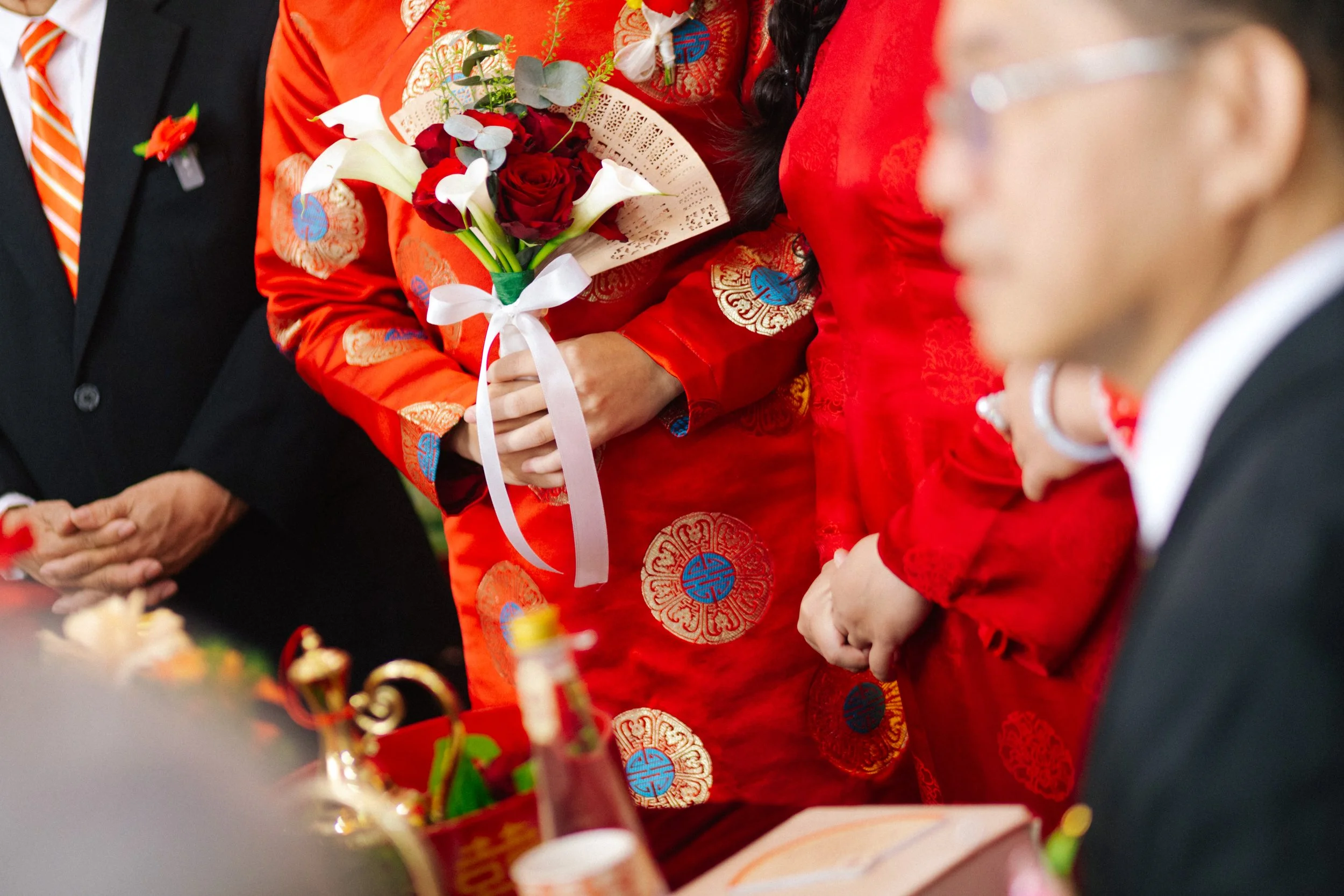People in traditional Chinese attire participating in a celebration, with one holding a bouquet of red and white flowers, and others dressed in formal suits.