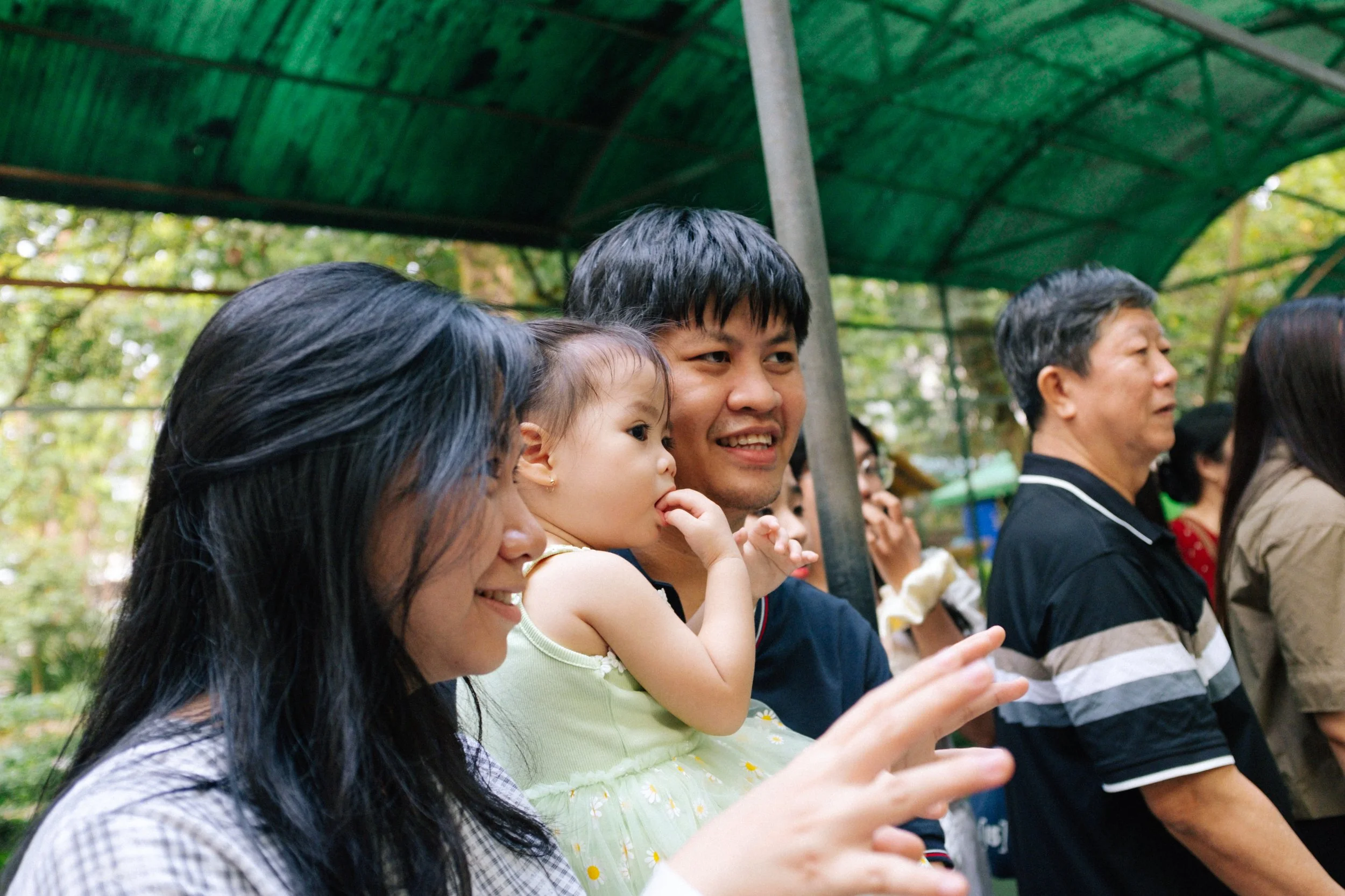 A group of people, including a woman, a man, a young girl, and an older man, standing outdoors under a green canopy, smiling and enjoying a moment together.