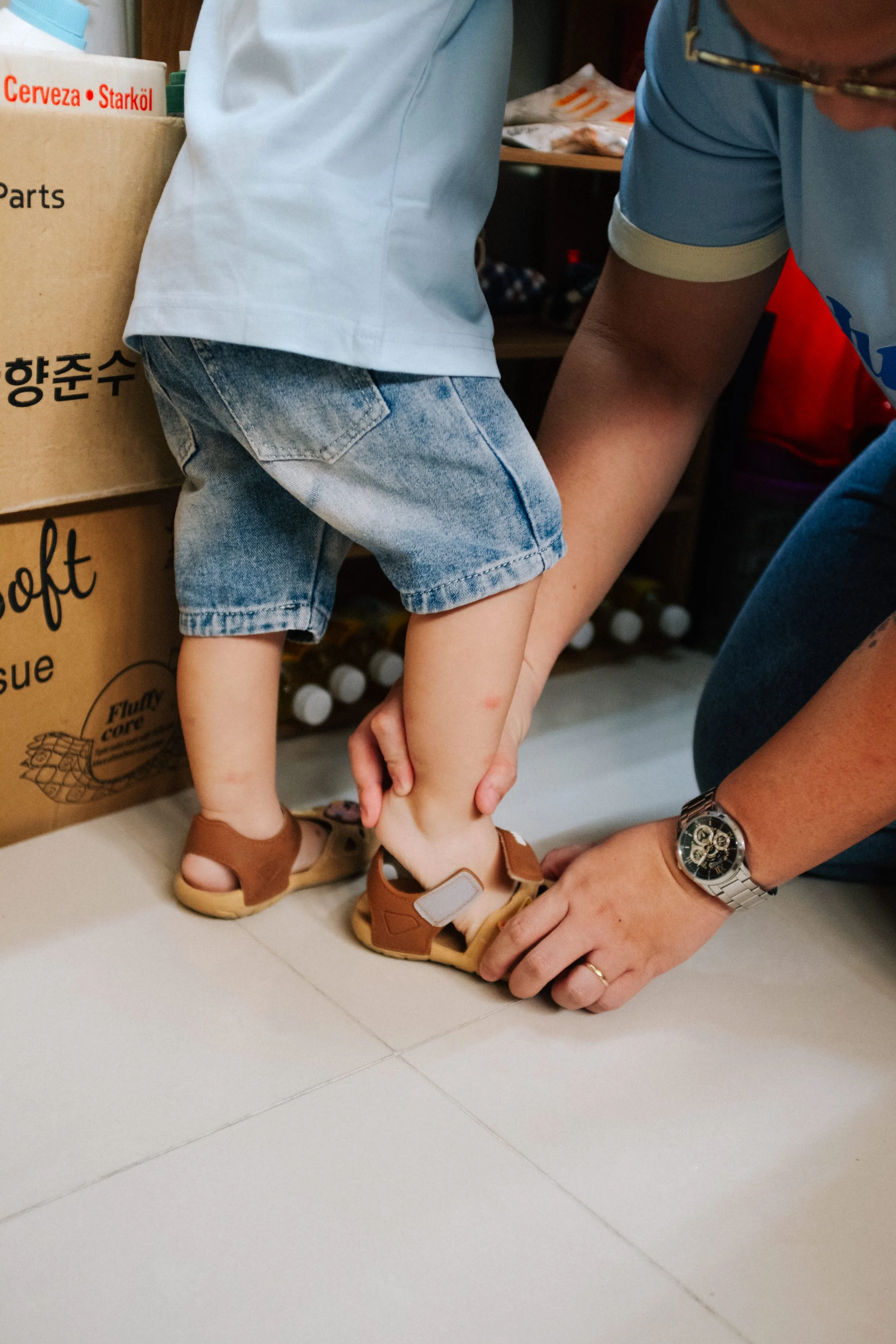 An adult helps a child put on sandals, with the adult holding the child's foot. The child is wearing denim shorts and a grey shirt, while the adult wears a blue shirt and a watch. There are boxes and shelves in the background.