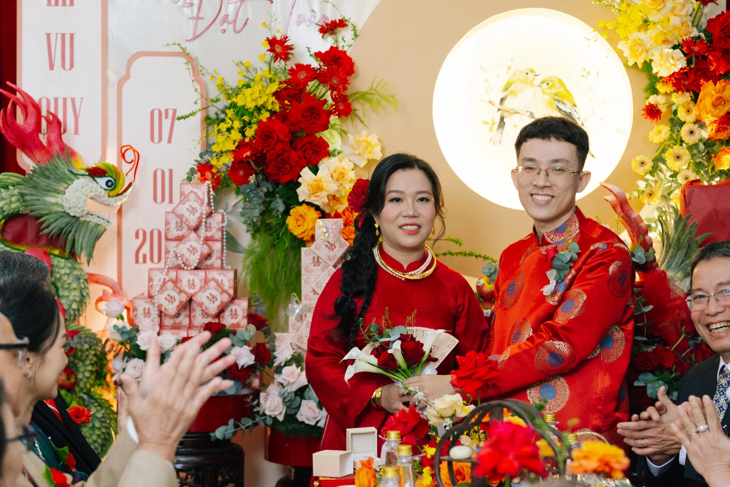 A couple dressed in traditional red attire at a wedding celebration, surrounded by vibrant flowers, a dragon decoration, and a round illuminated artwork of two birds.