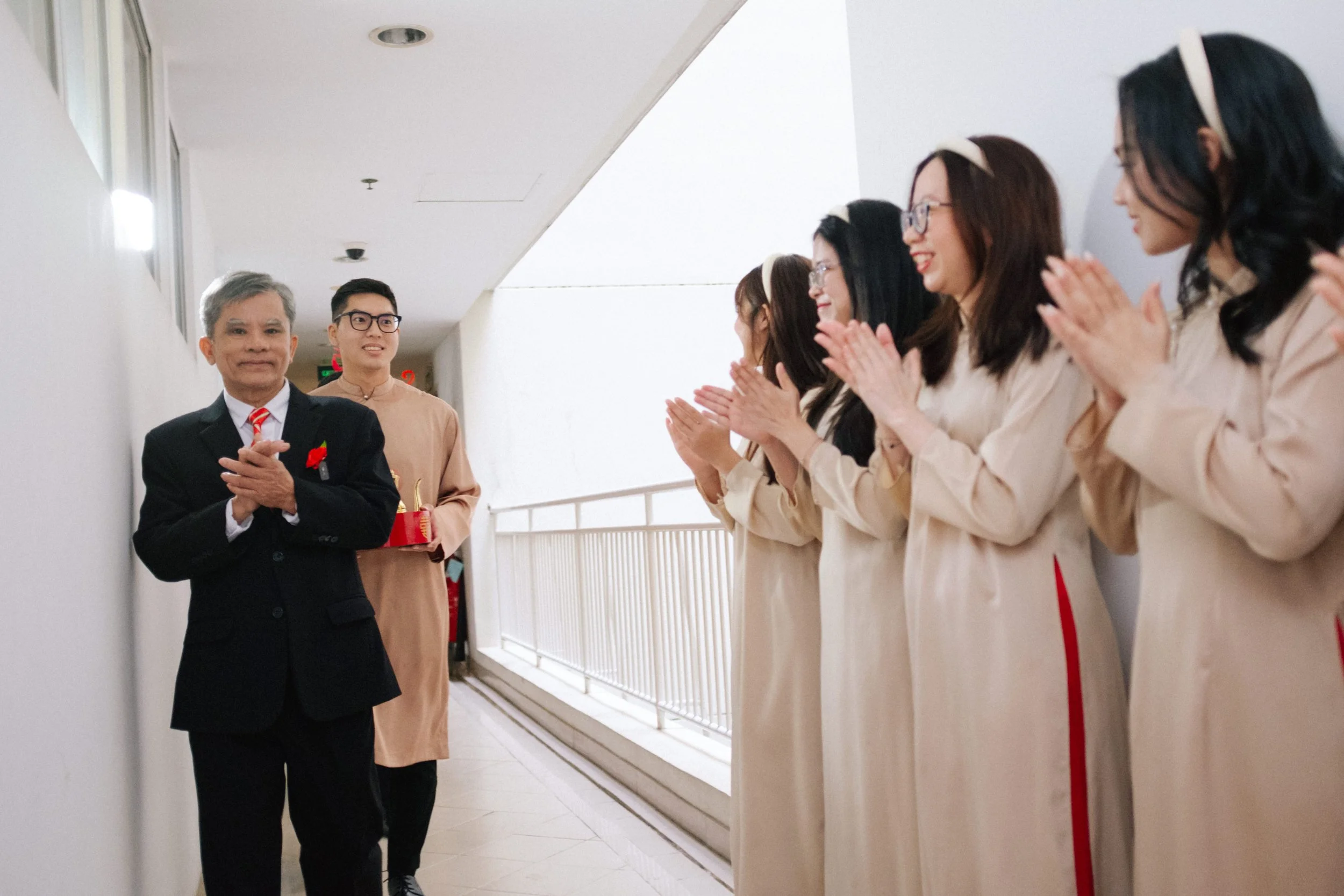 An elderly man in a black suit and red tie with a red flower pin stands in a corridor, clapping. Behind him, a young man in a beige traditional outfit is holding a red gift box. A group of women in beige dresses with red accents and headbands are cla