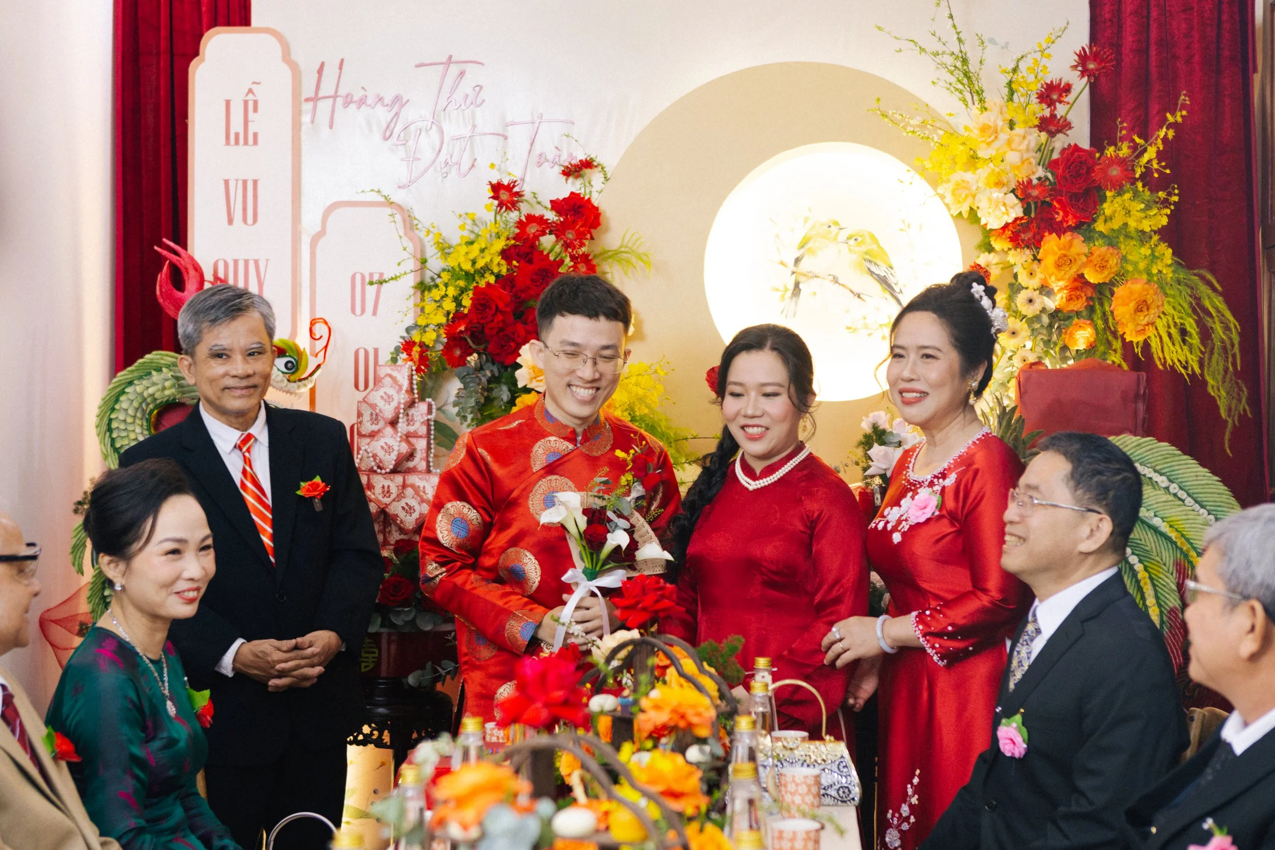 A Vietnamese wedding celebration with the bride and groom in traditional red áo dài, surrounded by family and friends in formal attire, with festive decorations and flowers.