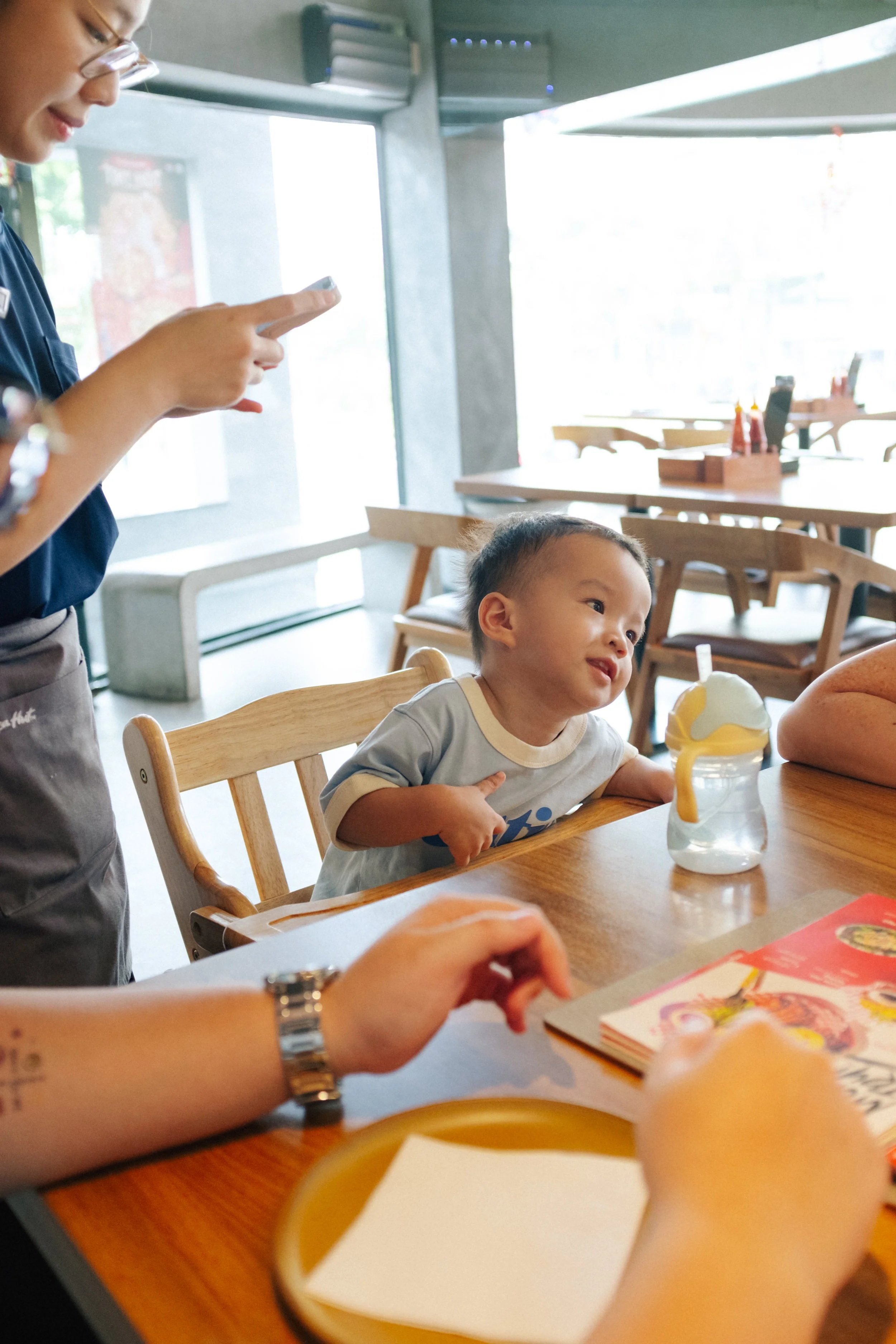 A young boy sitting at a wooden table in a restaurant, smiling and leaning toward the person sitting across from him, with a baby bottle with a lemon slice on top on the table. An adult with short hair and a wristwatch is reaching toward the table, a