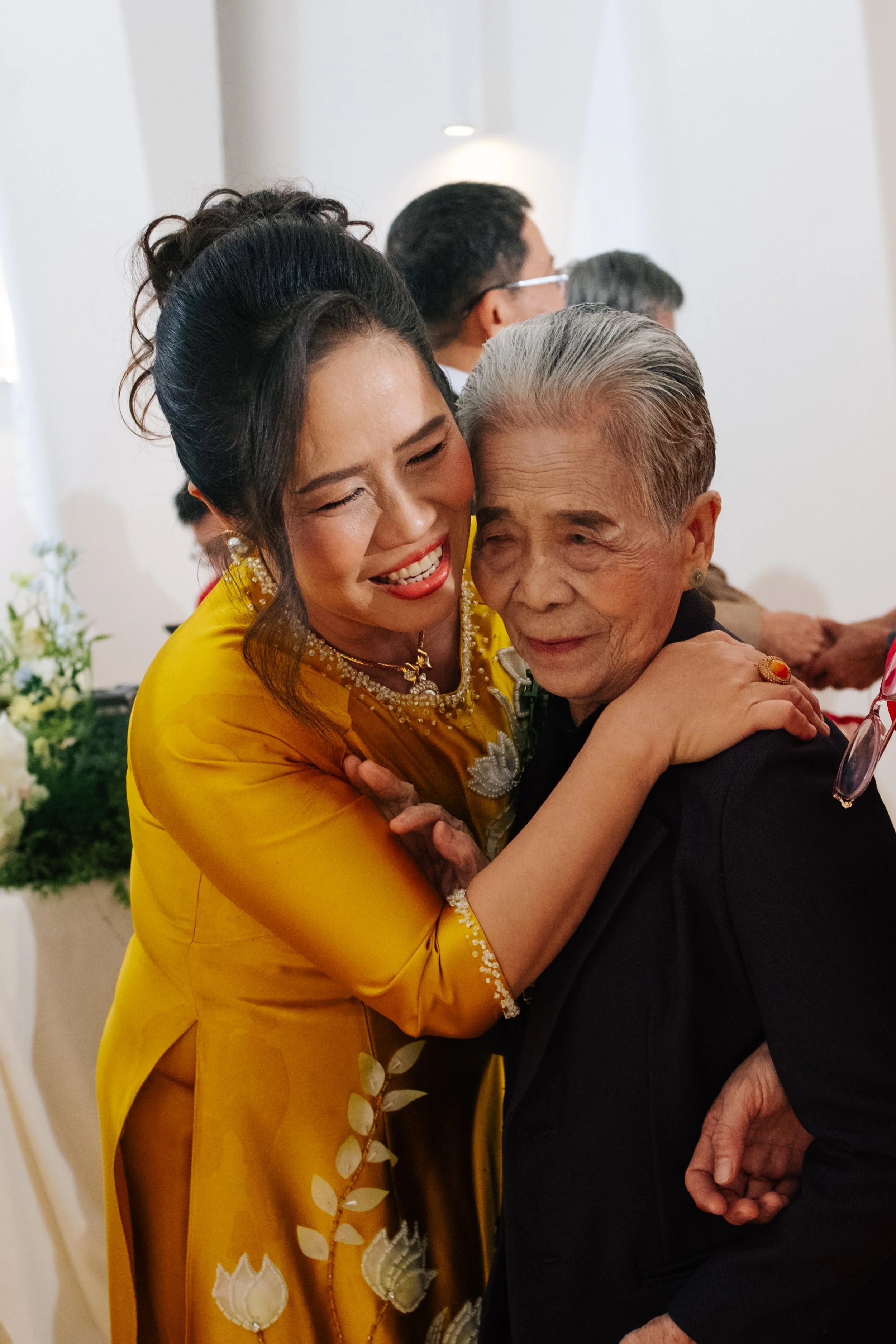 A woman in a yellow traditional dress hugging an elderly woman in a black dress at an indoor celebration or gathering.