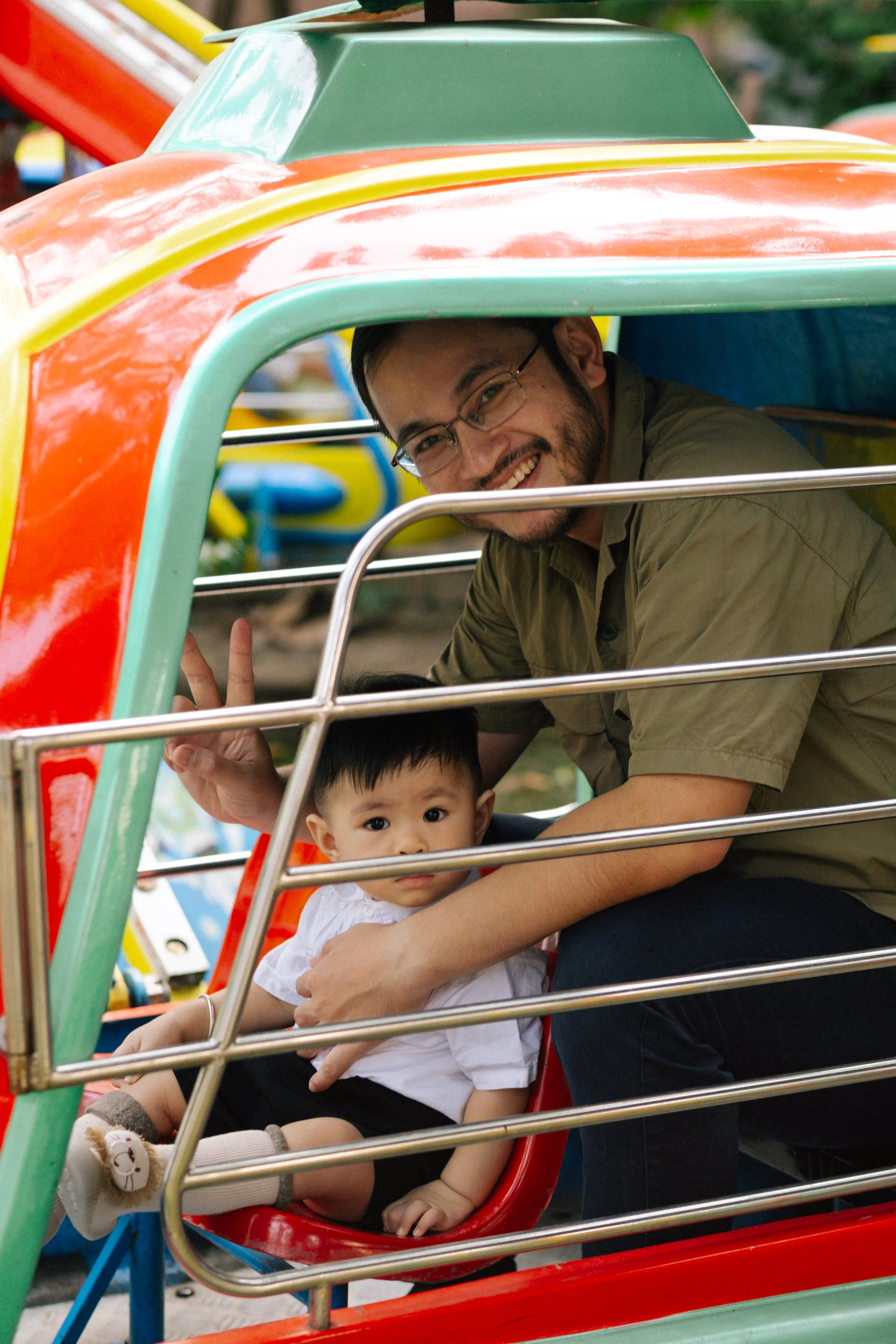 A man and a young boy sitting inside a colorful tram ride at an amusement park. The man is smiling and making a peace sign, while the boy looks curious.