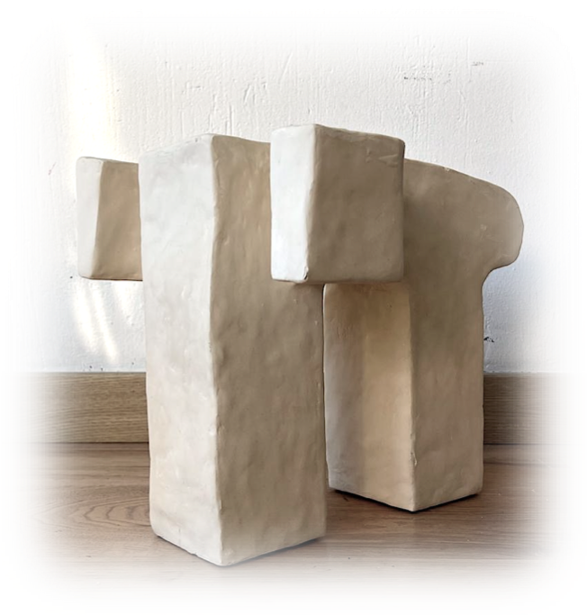 Escultura moderna de color beige, con formas geométricas y bloques, colocada sobre un suelo de madera contra pared blanca.