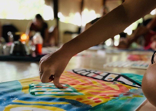 A person's hand touching a colorful fabric or artwork on a table with other people in the background.