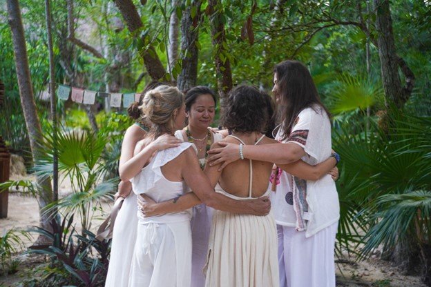 A group of five women standing outdoors in a circle with their arms around each other, sharing an emotional moment surrounded by trees and greenery.