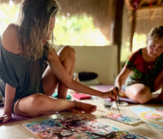 Three women sitting on the floor, painting colorful artwork together inside a rustic room.