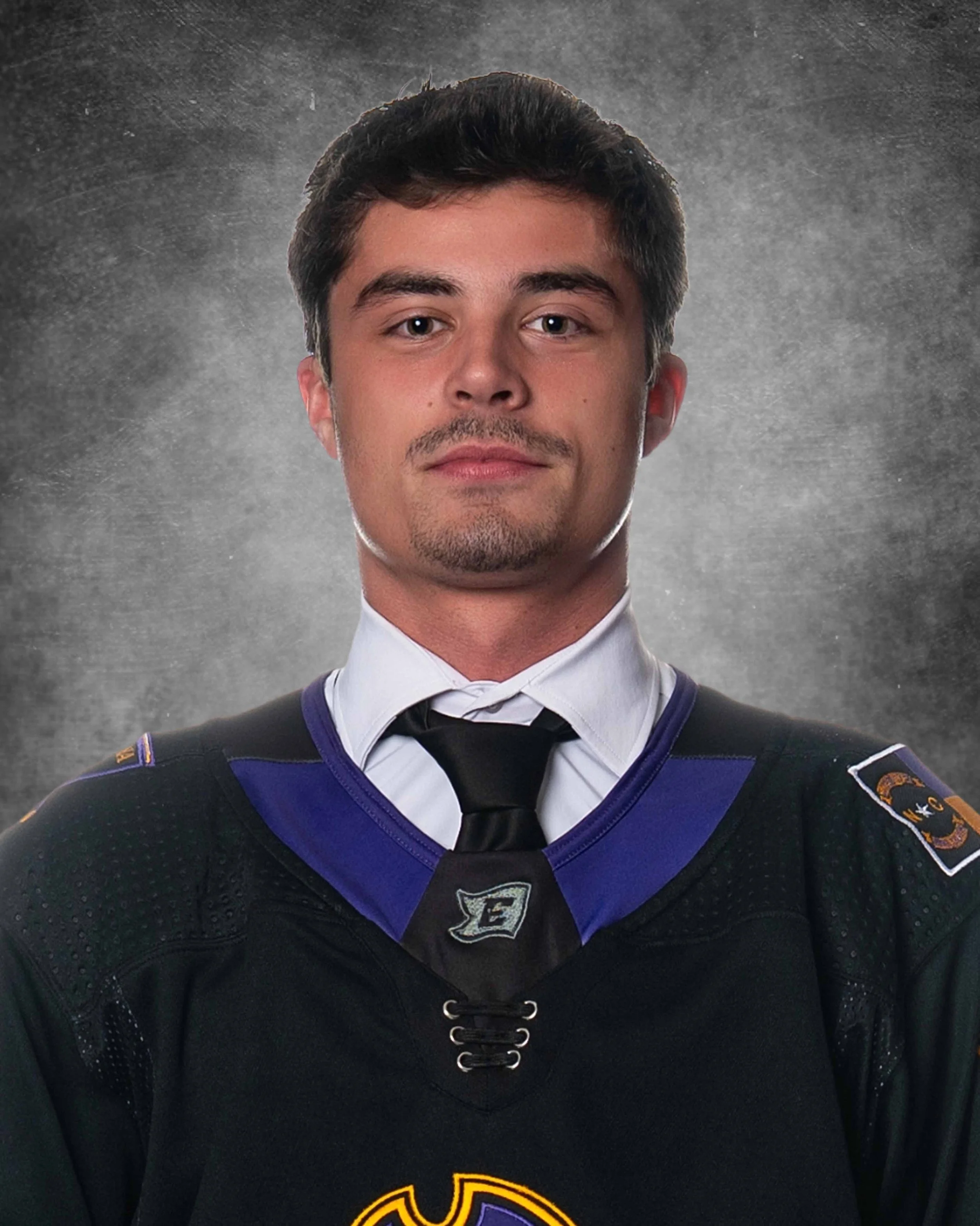 A young man wearing a black hockey jersey with purple and gold accents, a badge on the shoulder, and a black tie, against a textured gray background.
