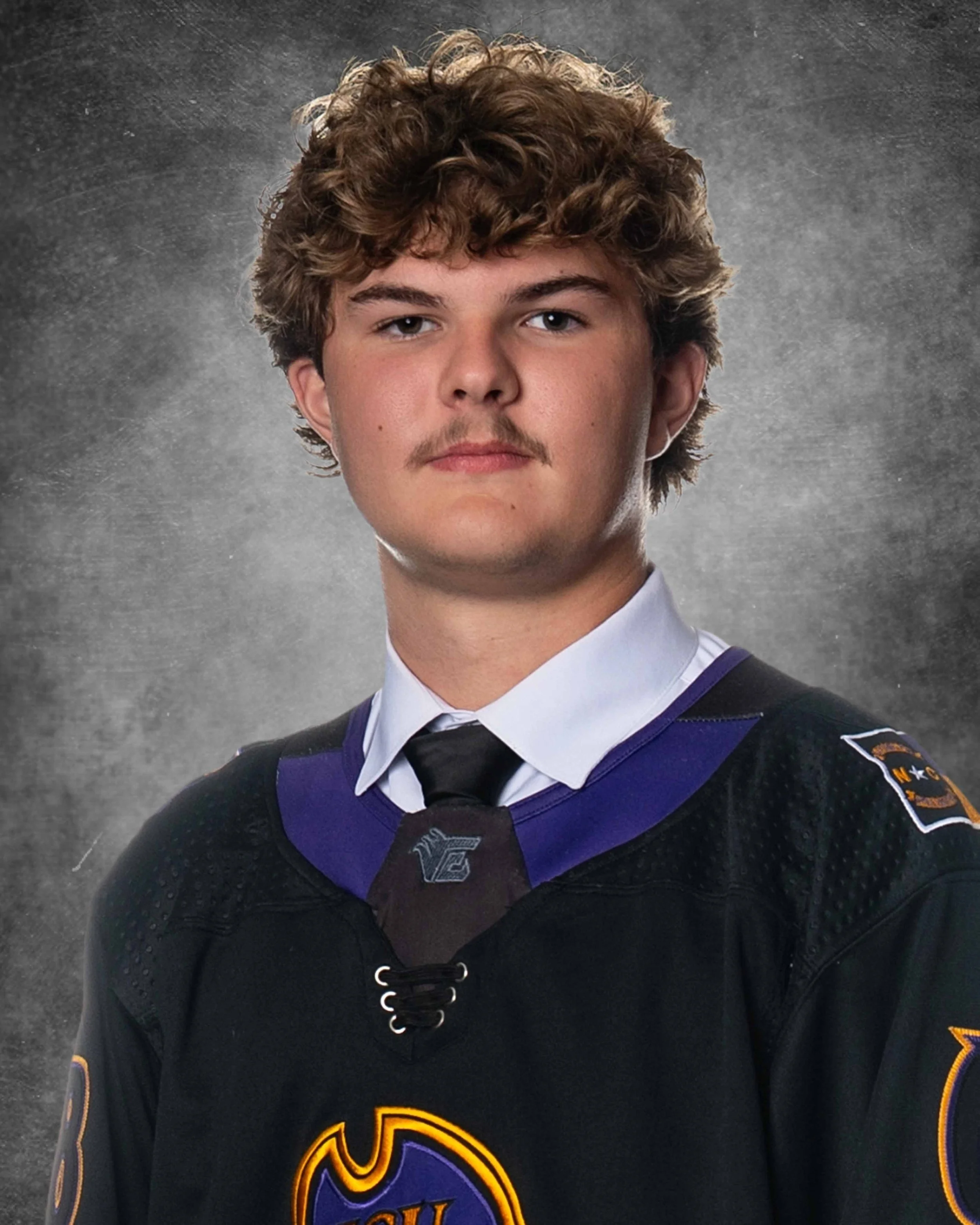 Young man with curly brown hair and light skin wearing a hockey jersey with purple, black, and orange colors, and a patch on the shoulder. He is looking directly at the camera with a neutral facial expression. The background is a textured gray gradie