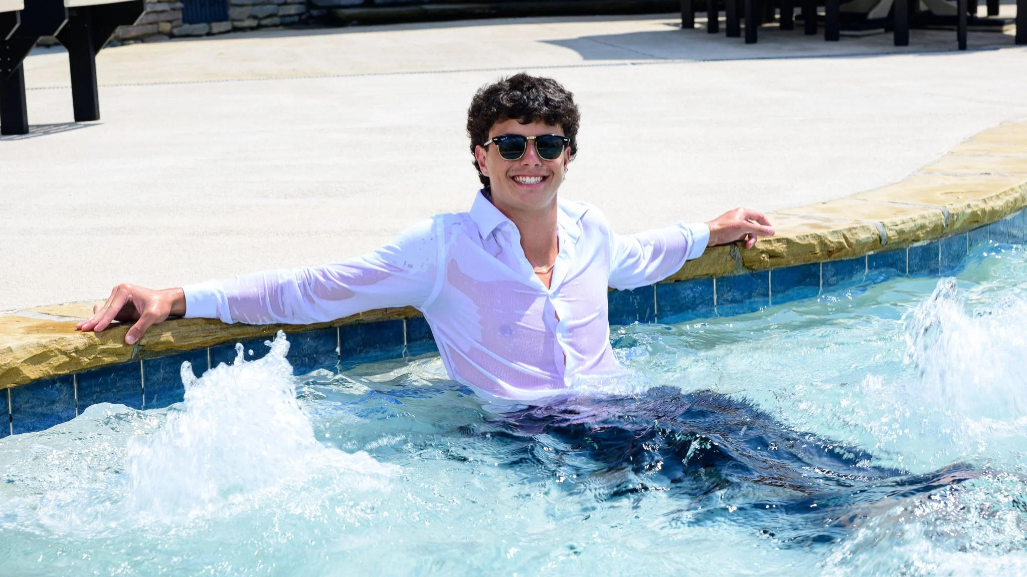 A young man with curly hair wearing sunglasses and a white shirt, smiling and relaxing in an outdoor swimming pool with his arms resting on the pool's edge on a sunny day.