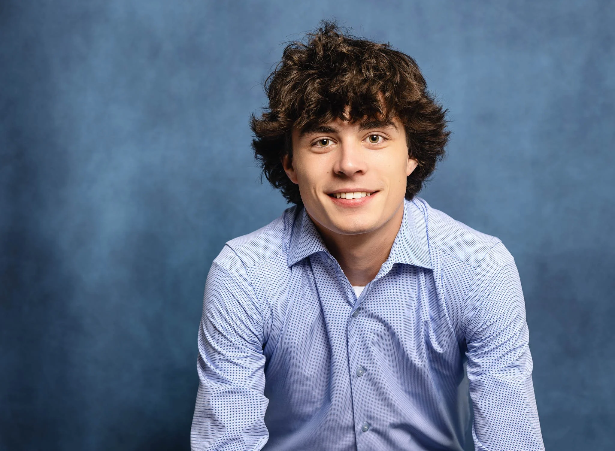 A young man with curly brown hair wearing a light blue collared shirt, smiling at the camera against a blue background.