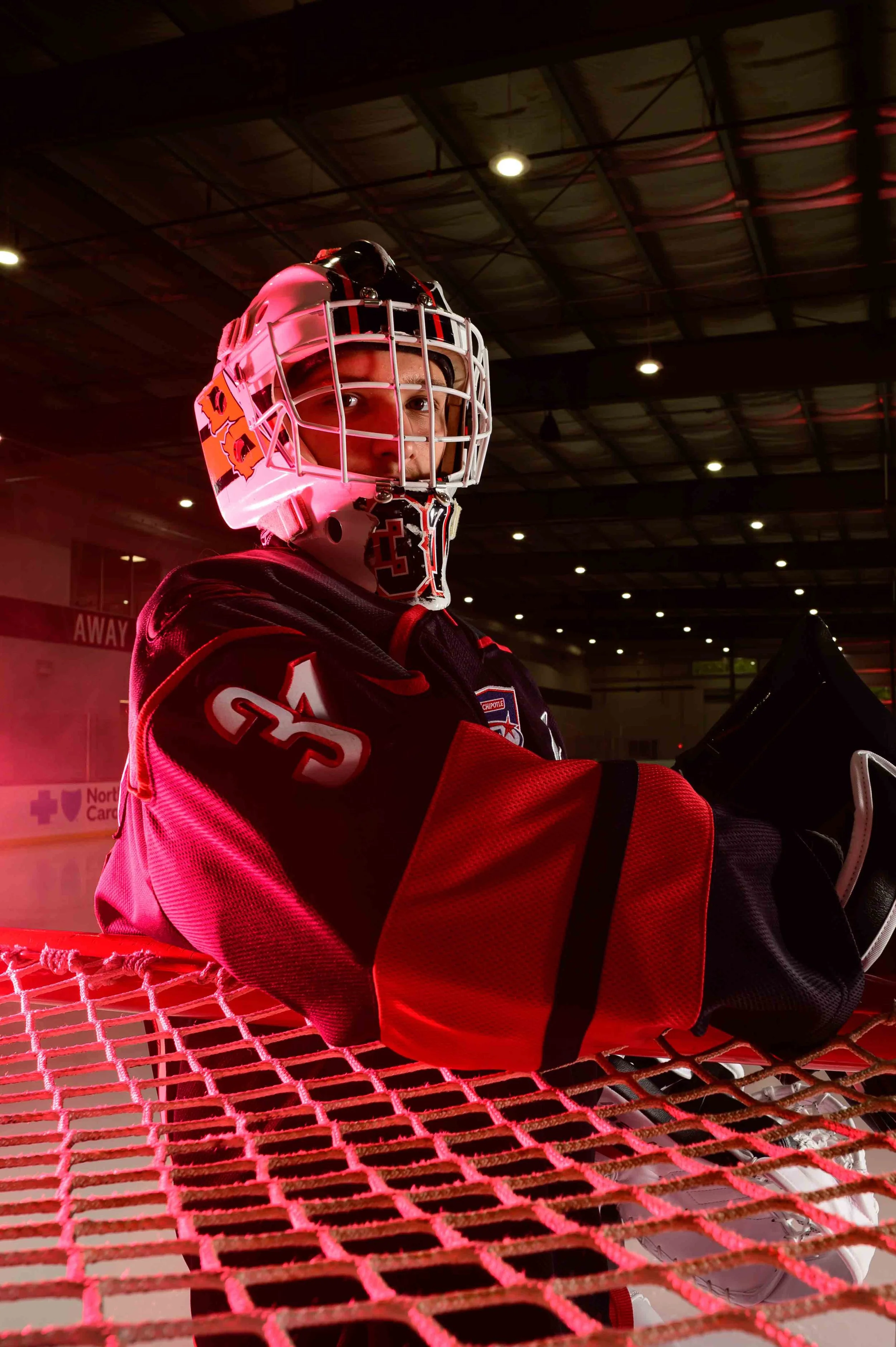 Ice hockey goaltender sitting on the bench in a rink, wearing a black, red, and white jersey and helmet, with his face partially visible through the helmet cage.