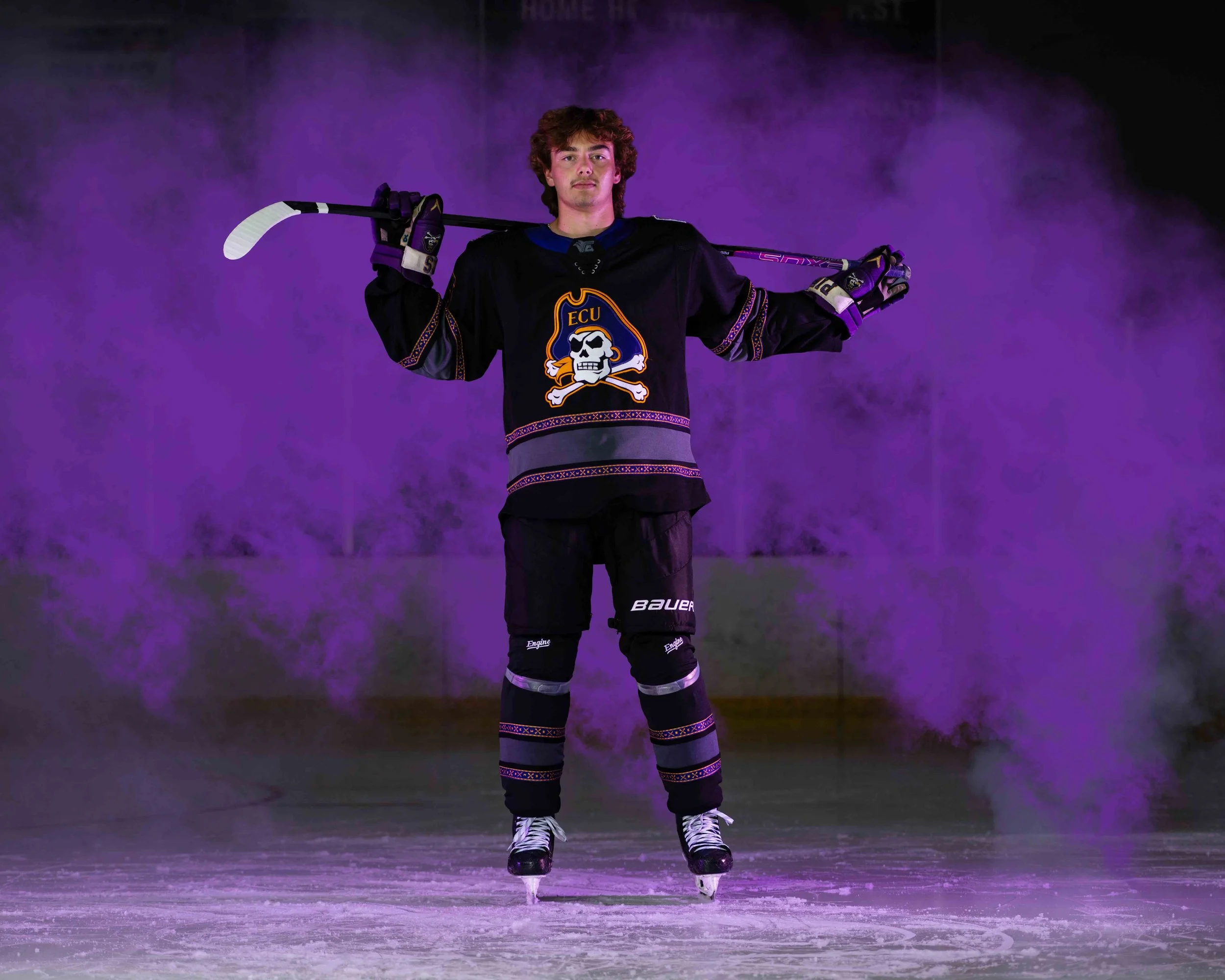 A young male ice hockey player stands on the ice rink, holding a hockey stick across his shoulders. He is wearing a black jersey with a skull and crossbones logo, black hockey pants, gloves, and skates, with purple and gold accents. The background is