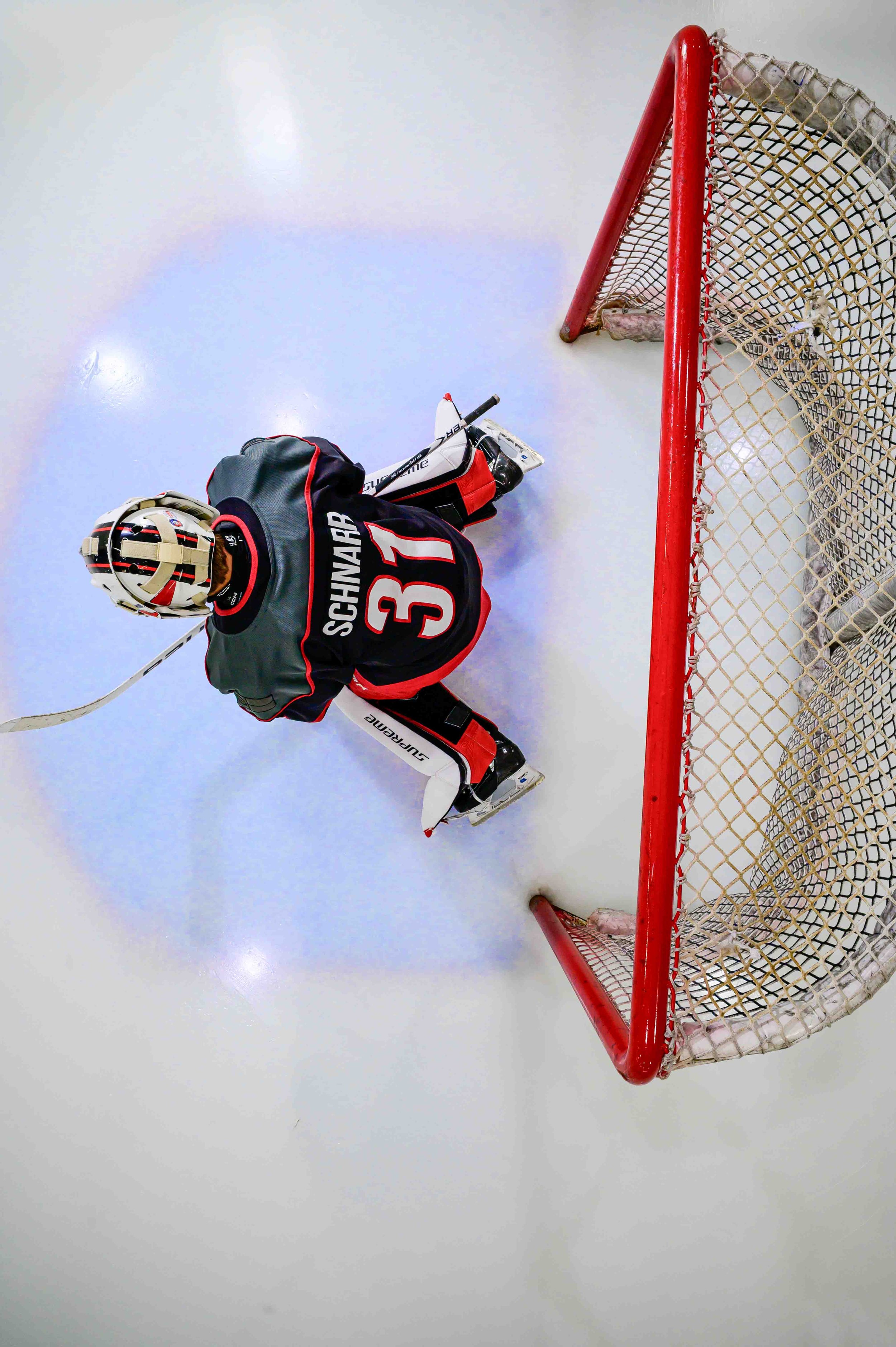 Hockey goalie in a crouched position in front of the goal, wearing full gear including a helmet, on ice rink.