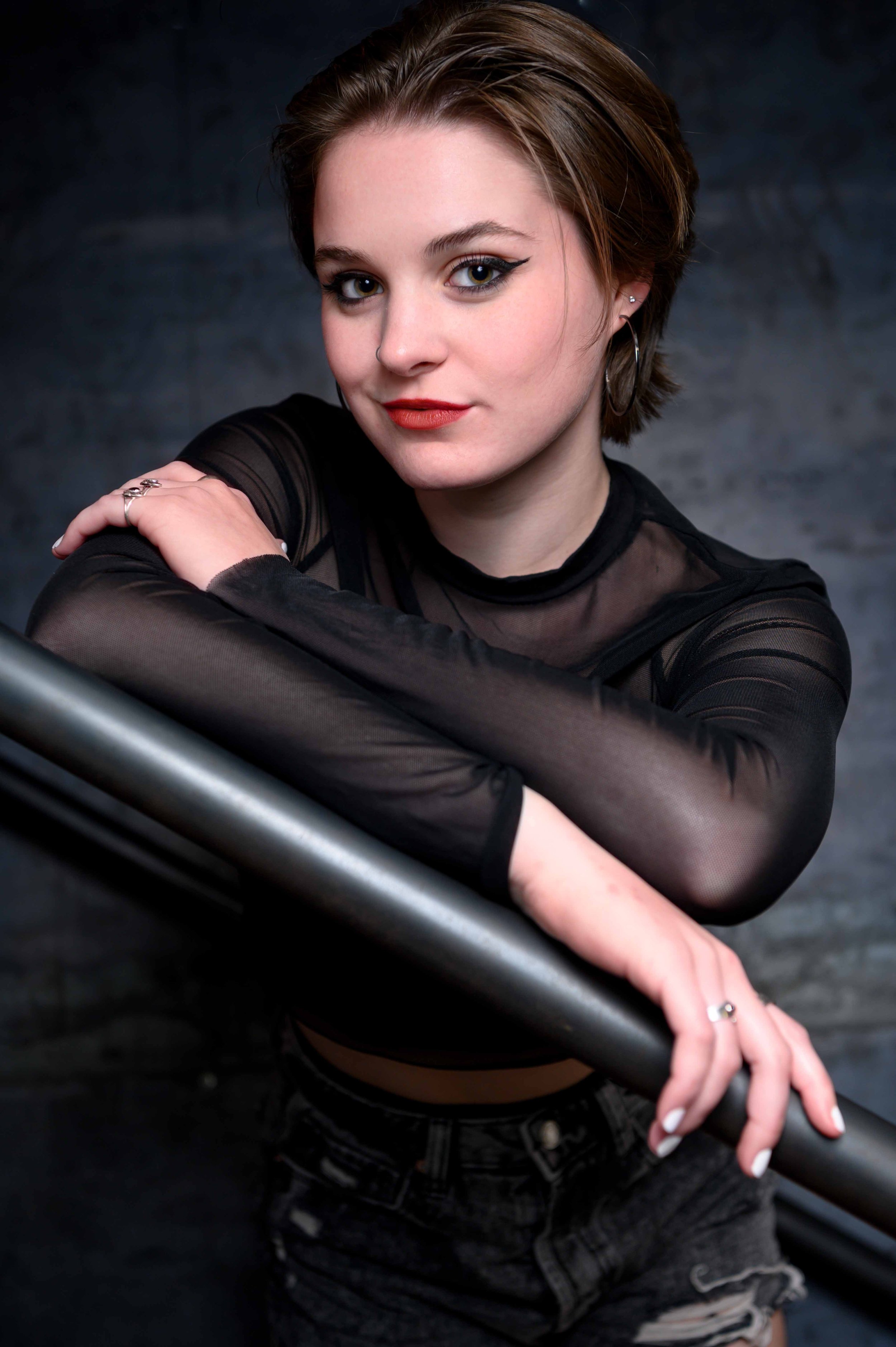 Young woman with short brown hair, wearing a black sheer top and black denim shorts, leaning on a metal railing against a dark background.