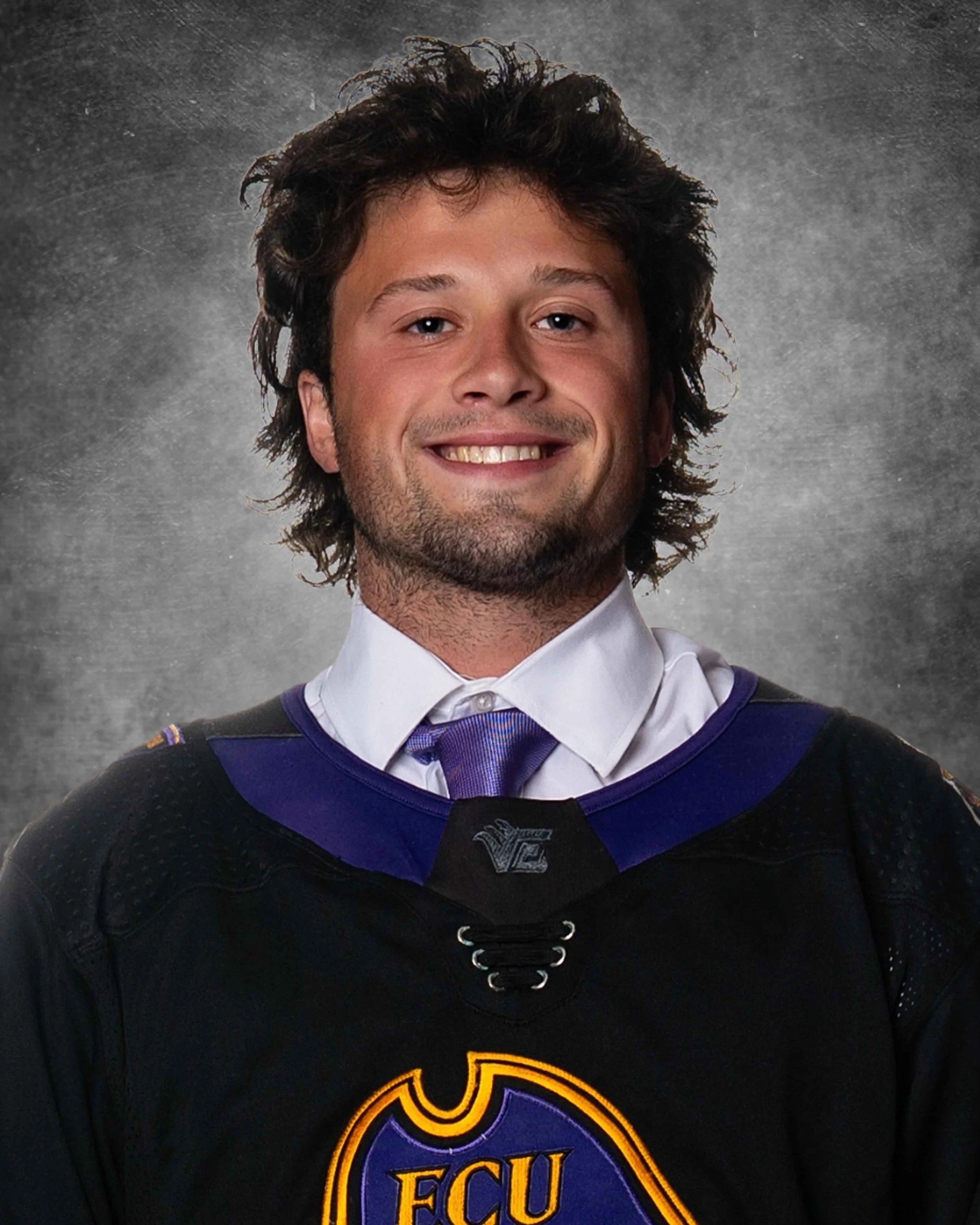 Close-up of a young man with brown curly hair, wearing a black hockey jersey with purple and yellow accents, and a white dress shirt underneath, smiling at the camera against a gray textured background.