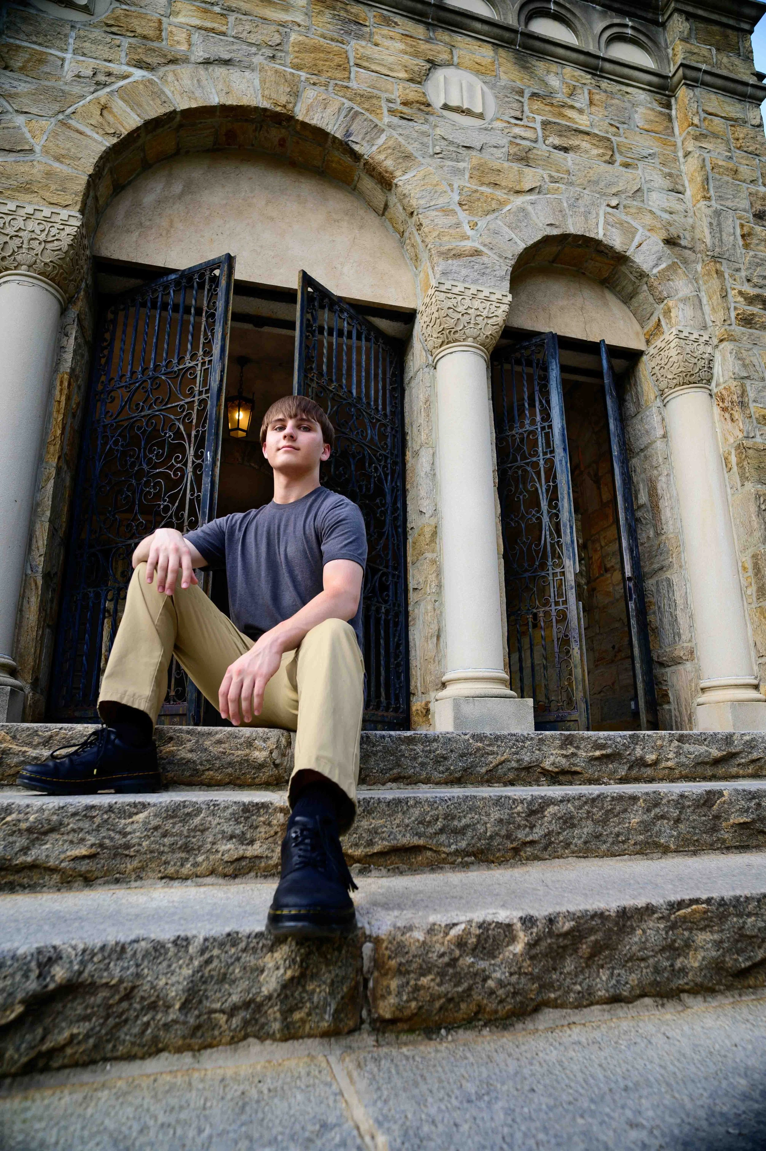 A young man in a gray t-shirt and khaki pants sitting on stone steps in front of an ornate wrought-iron gate and stone building with columns.