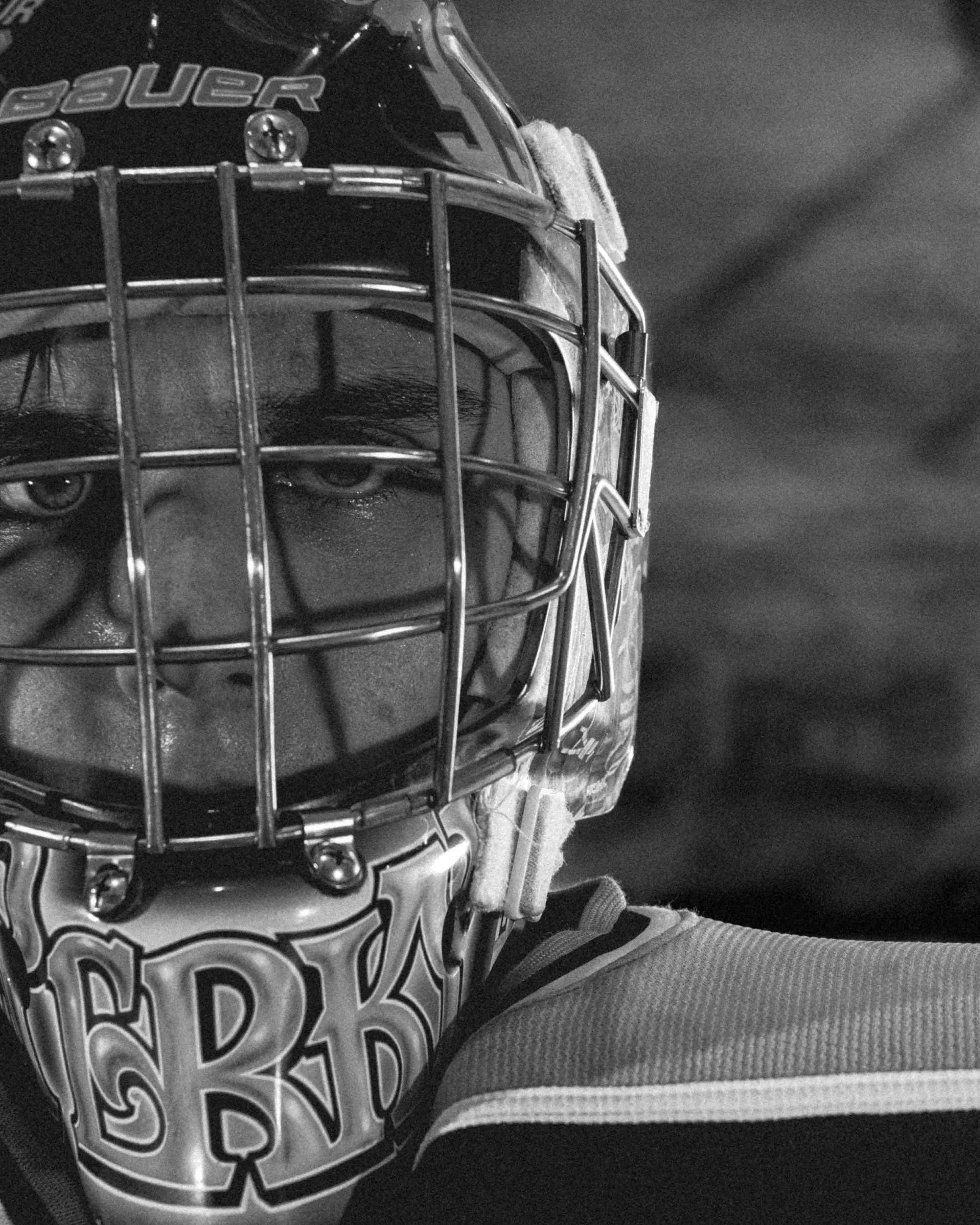 Close-up of a hockey goalie wearing a mask with a cage and a helmet, looking directly at the camera in black and white.