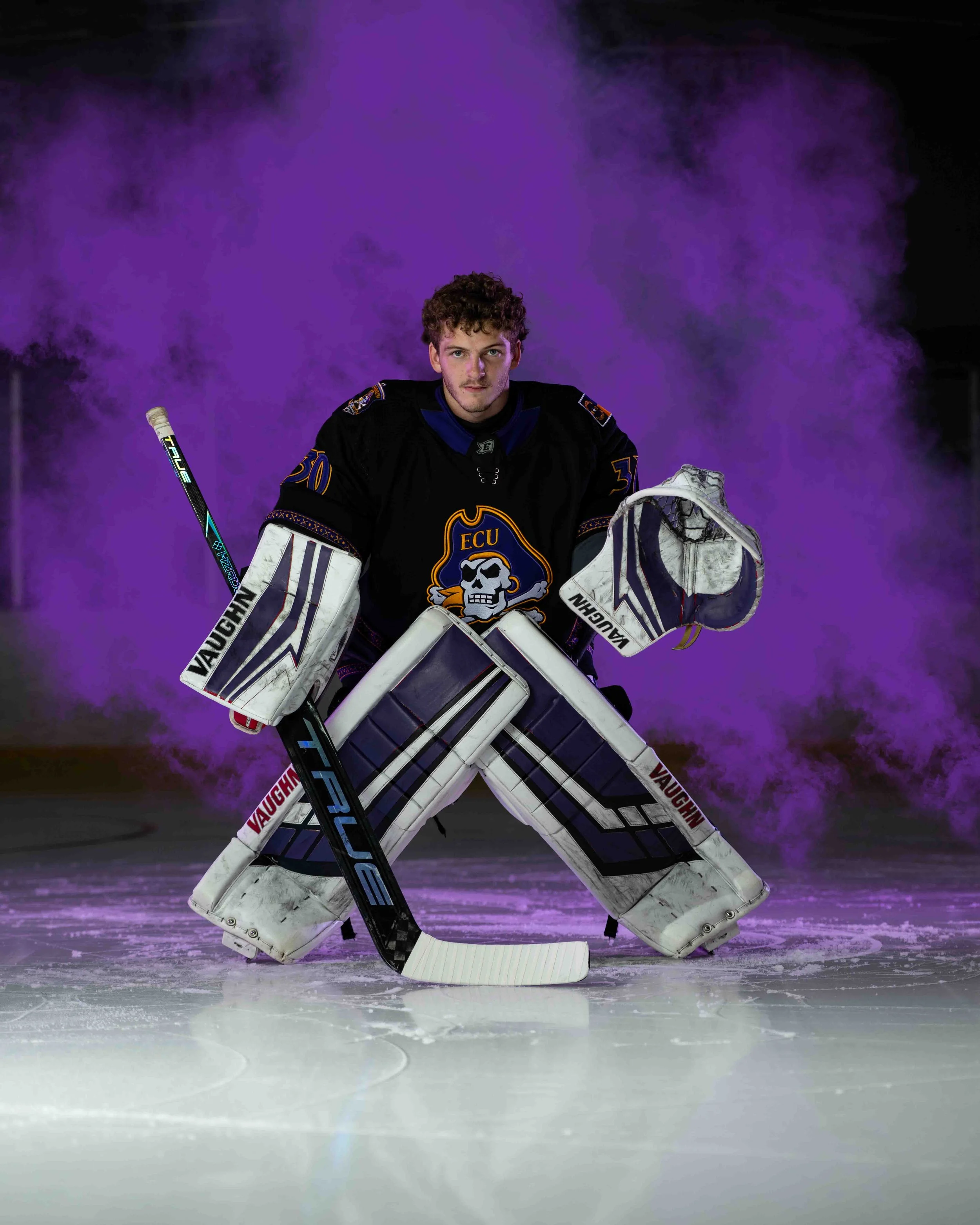 An ice hockey goalie in black jersey with a skull logo, standing in front of a purple fog or smoke, on an ice rink.