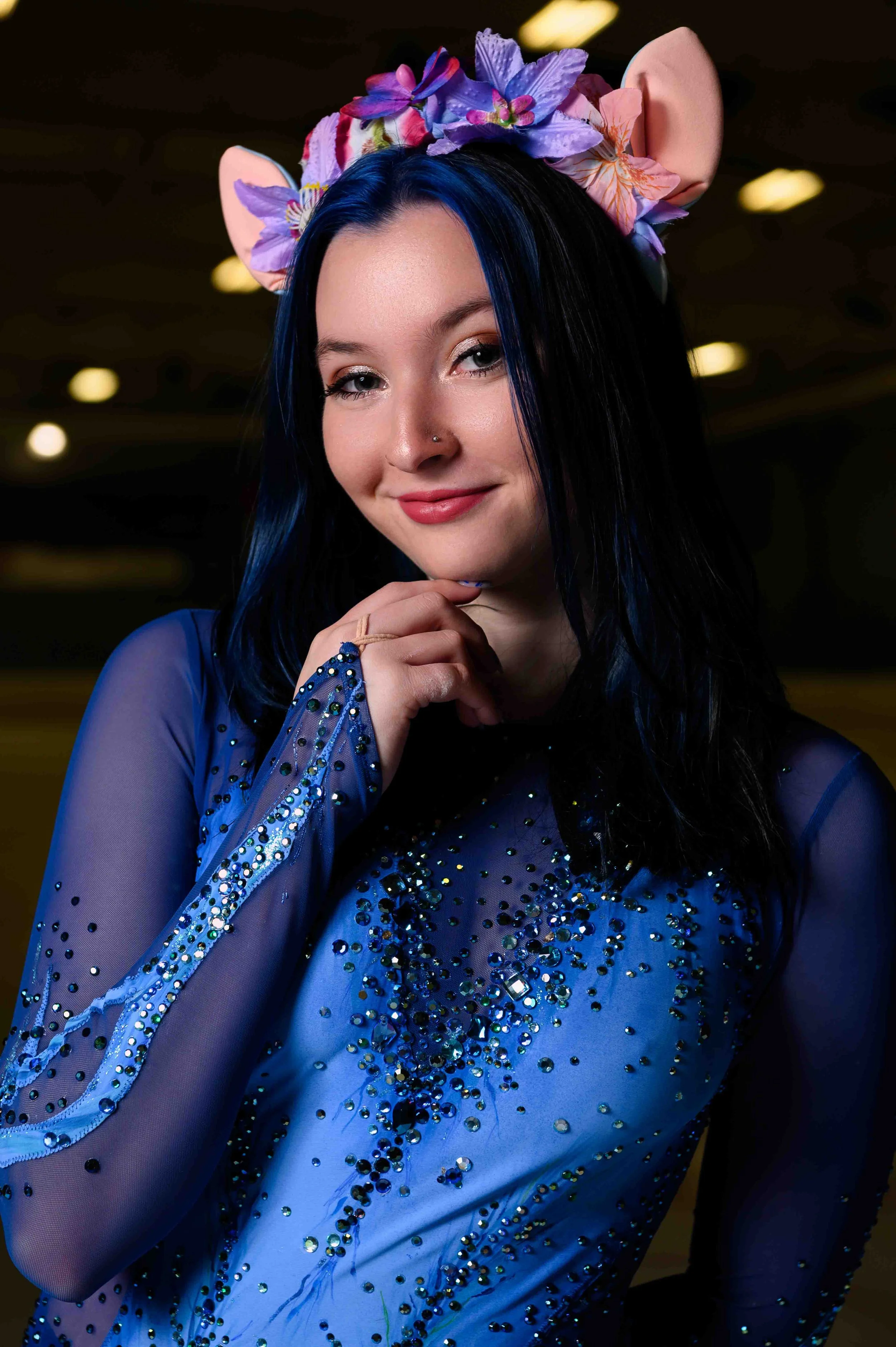 A young woman with long dark hair and a nose piercing is wearing a blue, sequined, sheer top and a headpiece with pink, purple, and blue flowers and light pink ears, posing with her hand near her chin and smiling in a dimly lit environment.