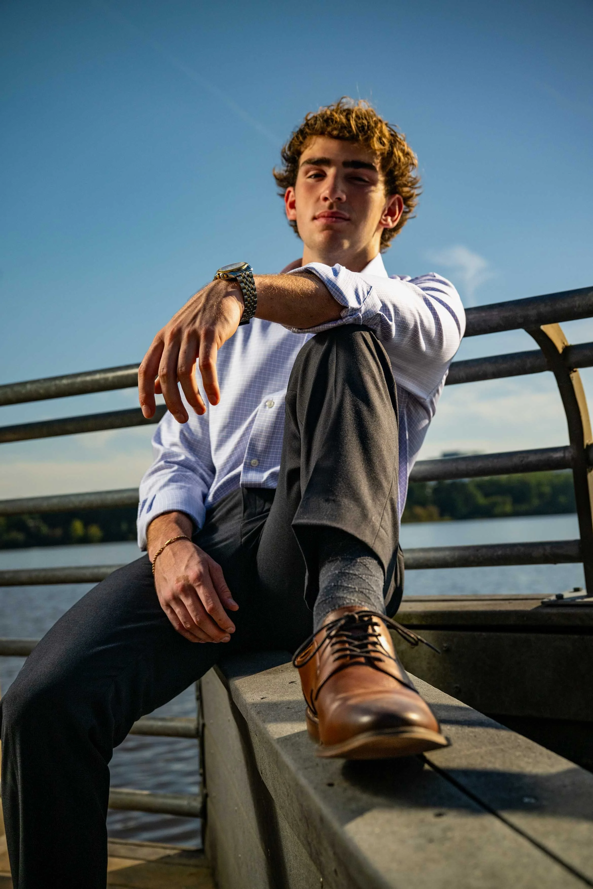 A young man with curly hair sitting on a dock by the water, wearing a light blue button-up shirt, dark pants, and brown leather shoes, looking confidently at the camera with a clear sky and distant trees in the background.