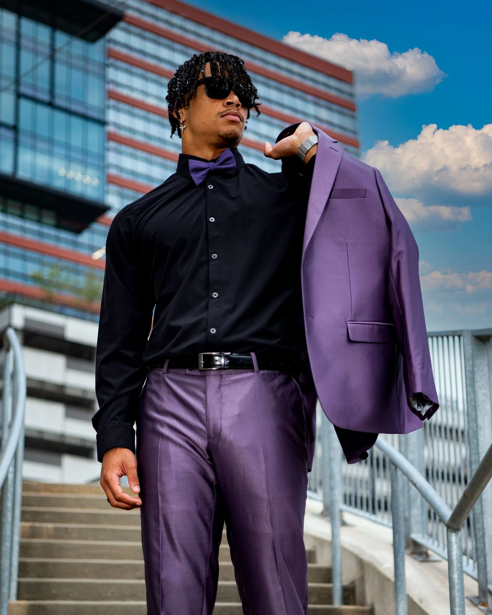 A stylish man dressed in black shirt with purple pants and matching purple blazer, holding the blazer over his shoulder, standing outdoors in an urban setting with stairs, metal railing, and modern glass building behind him.